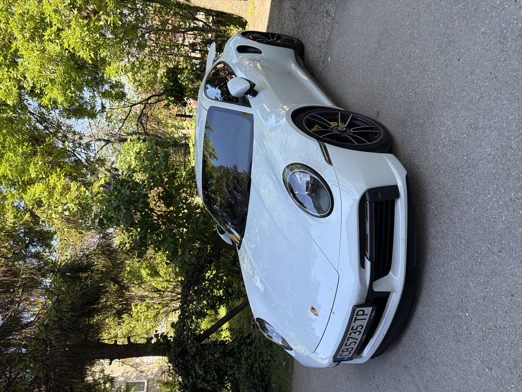 A white Porsche 911, displaying the license plate "CB 5735 TP," is parked on a gray asphalt road. The location appears to be in a tree-lined area, likely a residential street in Pleven, Bulgaria. The car is situated near vegetation, including trees and ivy. There are no people visible in the scene.