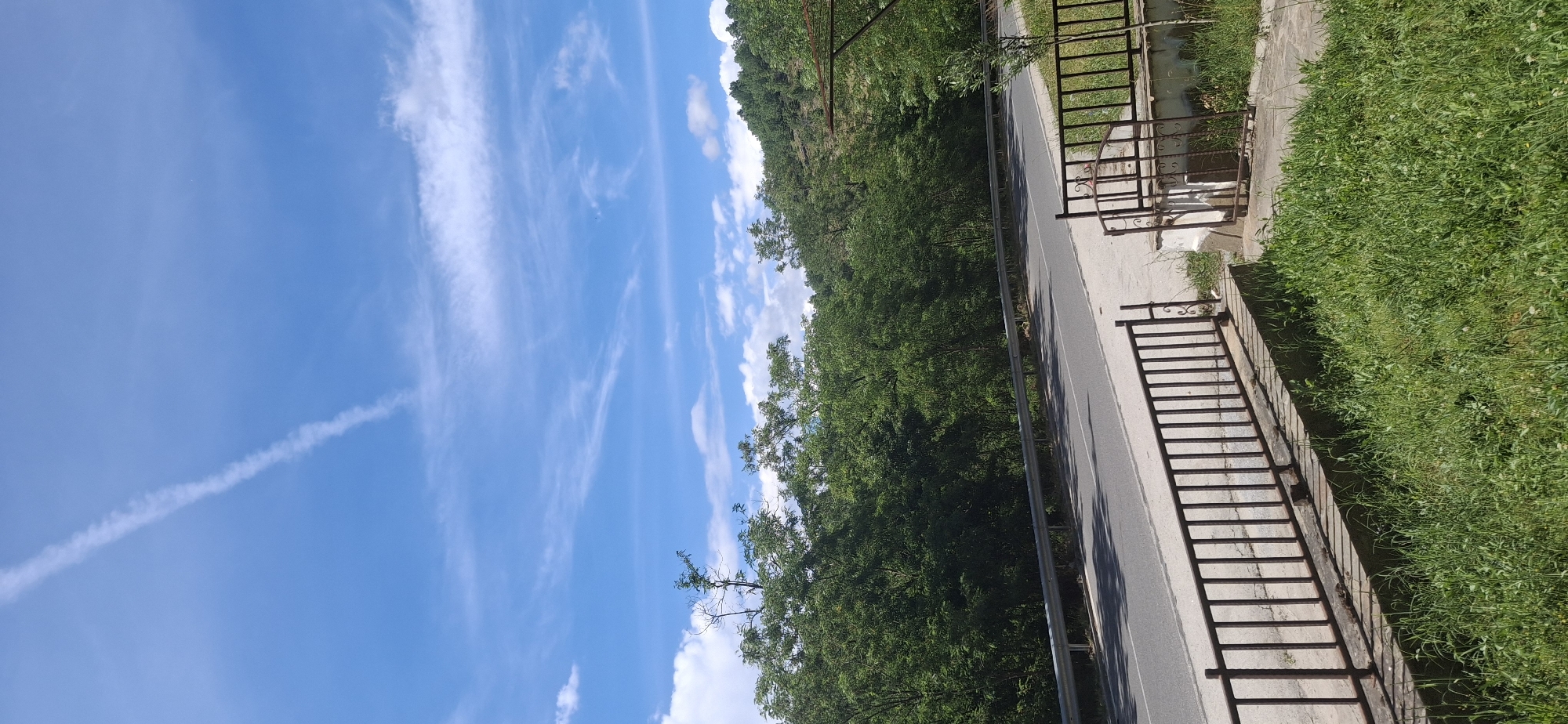 This is a vertical, eye-level, outdoor photograph of a road winding through a green, hilly landscape under a bright blue sky with wispy white clouds. In the foreground, there is a metal fence with vertical bars and some decorative scrollwork on the right side. Beyond the fence is a concrete walkway and a patch of green grass. A paved road runs parallel to the fence, separated by a silver metal guardrail. The road curves to the left and disappears into a dense forest of green trees. The trees cover the slopes of the hills, which rise in the background. A thin contrail stretches across the sky from the upper left to the lower right. The scene appears to be a rural or semi-rural area.