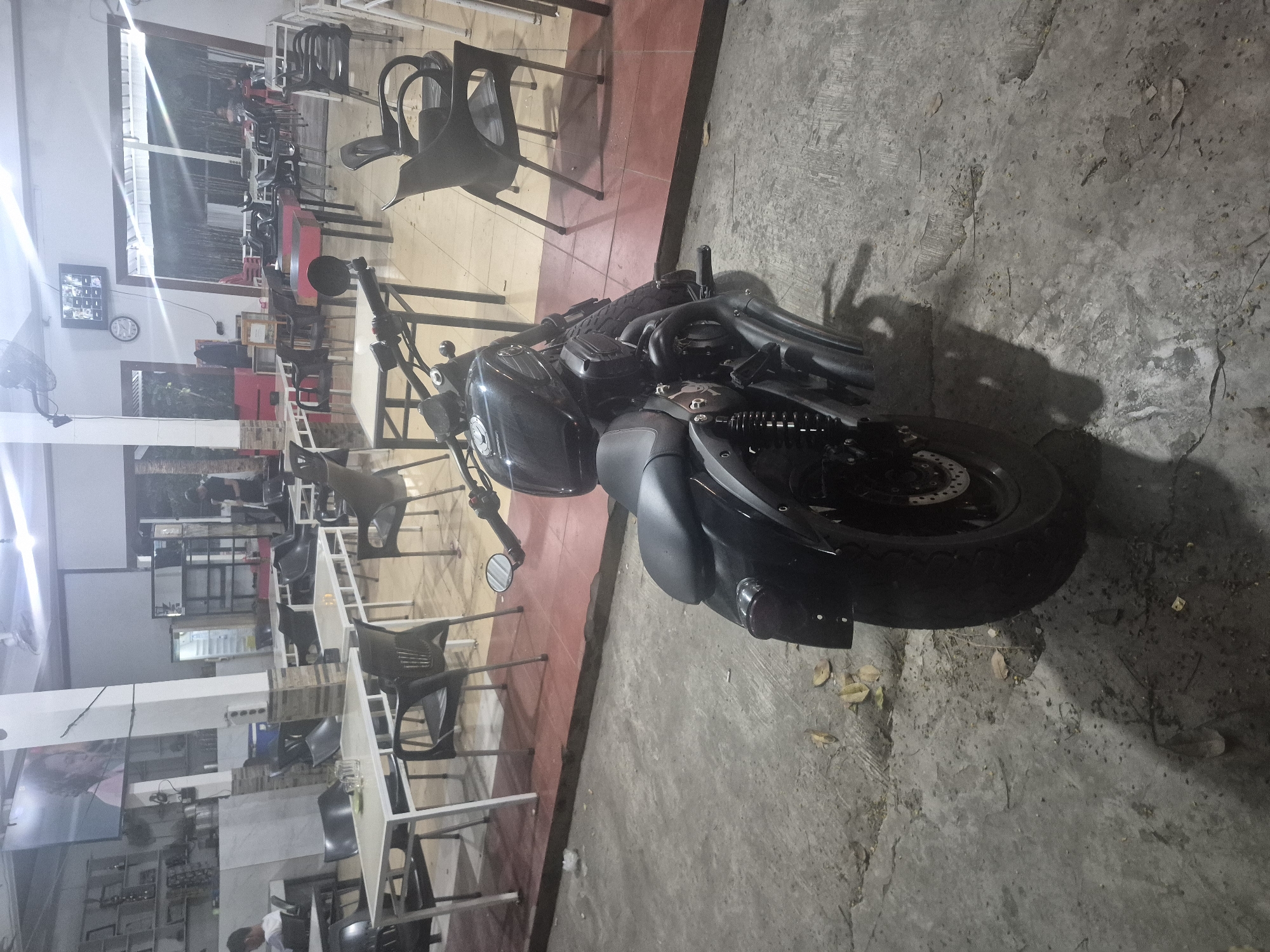 The image shows a black motorcycle parked on a concrete surface, possibly a sidewalk or parking area, in what appears to be an outdoor section of a restaurant or cafe. Tables and chairs, mostly empty, are visible inside the building behind the motorcycle. The scene is generally dimly lit, suggesting it is either nighttime or a poorly lit area.
While it's possible the image was taken in Lhokseumawe, Indonesia, based on the setting and context, this cannot be confirmed.