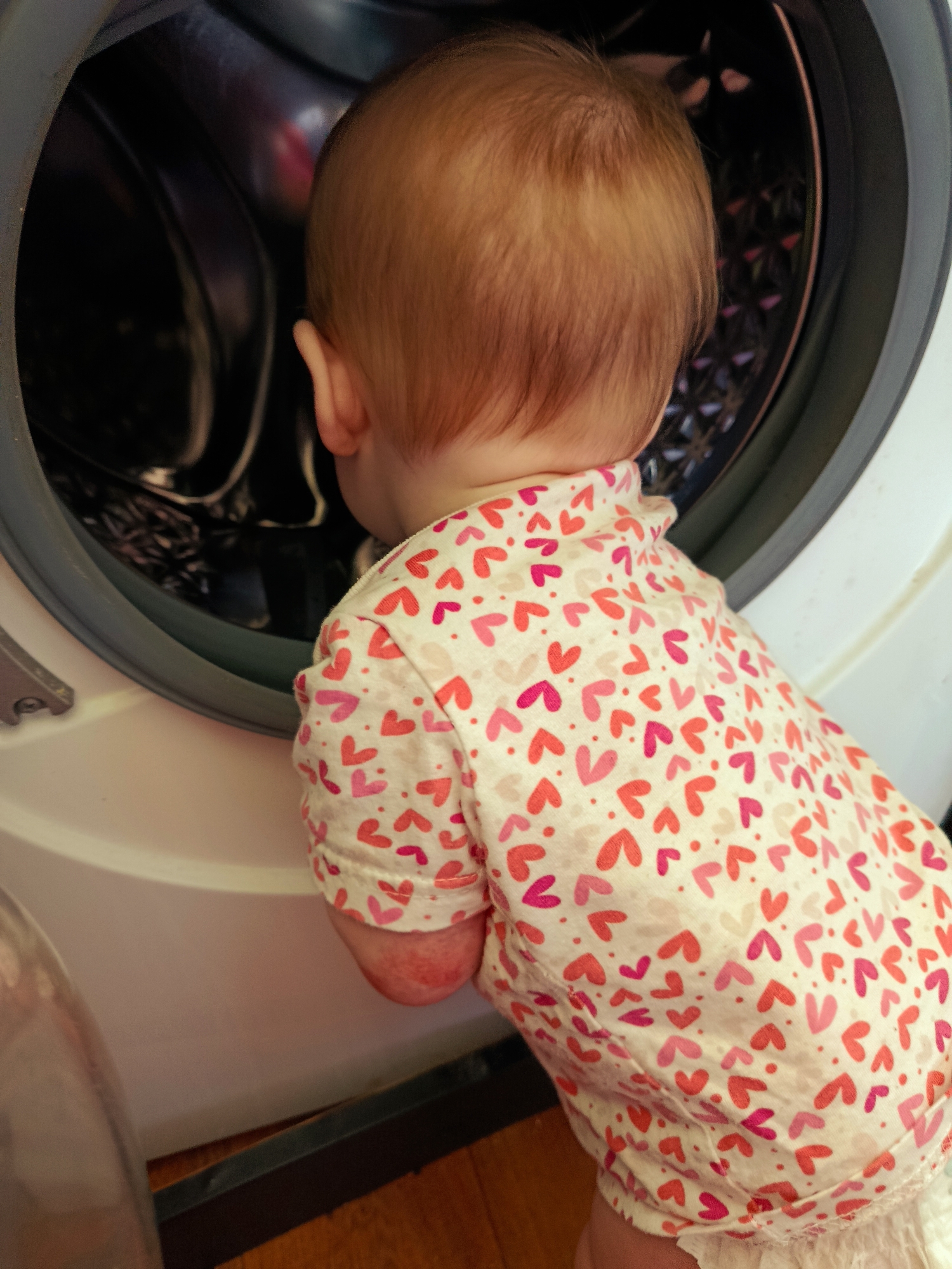 A baby is peering into the open door of a front-load washing machine. The baby has light, reddish-blonde hair and is wearing a white shirt patterned with pink and red hearts. The washing machine is white with a dark gray door frame. The interior of the machine appears to be stainless steel. The background consists of a wooden floor and part of another appliance, possibly a refrigerator. The location in the city of Фоки, Russia, is not discernible from the image.
