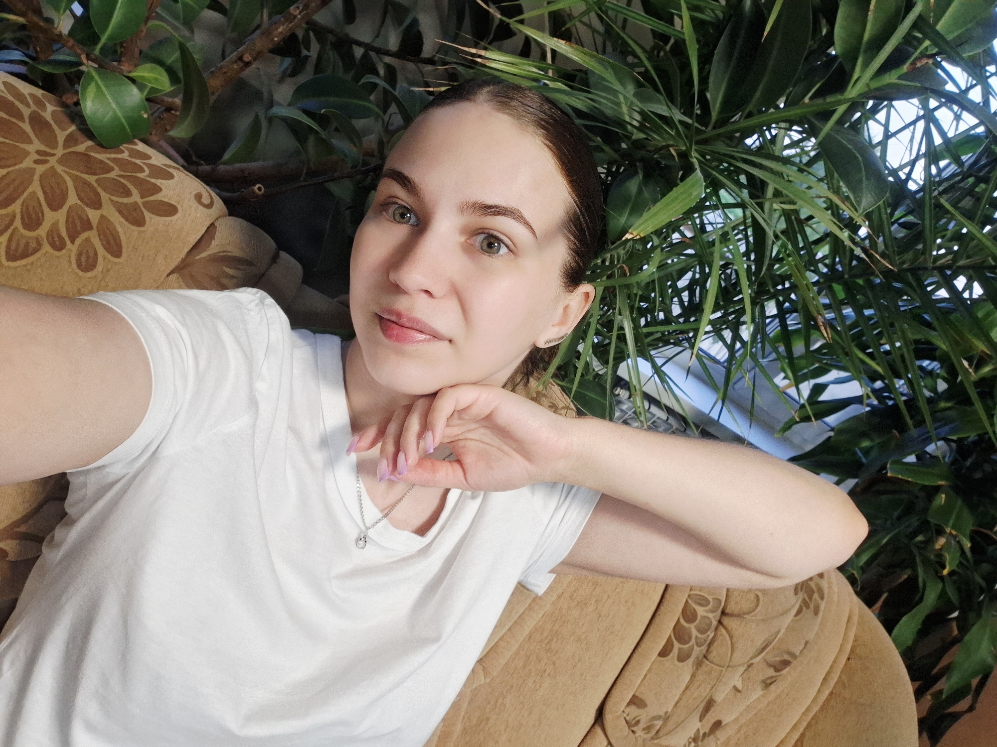 A woman is posed in a selfie-style photograph. She is light-skinned with brown hair pulled back and green eyes. She is wearing a white T-shirt and a delicate silver necklace with a small pendant. Her left hand is raised and resting on her cheek, with manicured nails painted a light pink.
The woman is positioned on a brown floral patterned sofa. Behind her are two types of green plants. One appears to be leafy with rounded edges, while the other has slender, blade-like leaves.
Based on the presence of Russian toponyms (Nizhnyaya Gar' is a place name in Russia), the location is likely somewhere within Russia. However, the specific city or environment of Nizhnyaya Gar' cannot be identified from the information provided in the image.