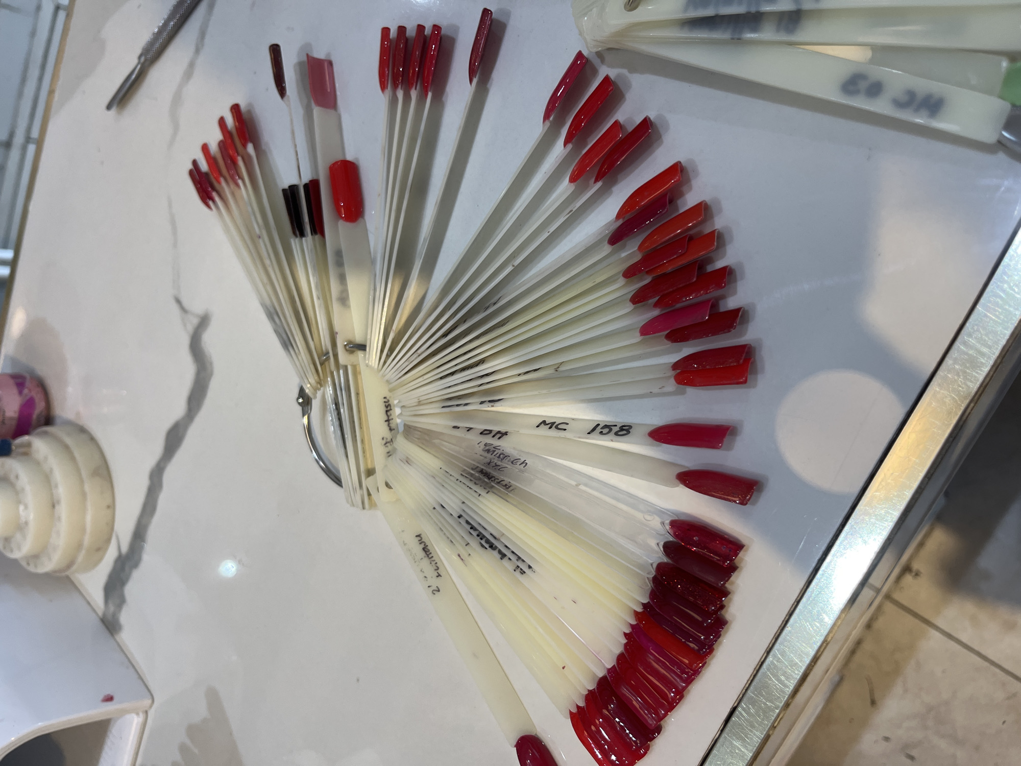 The image shows a collection of nail polish samples displayed on white plastic sticks arranged in a fan shape, presumably in a salon or cosmetic setting. The samples primarily feature various shades of red, from light pinkish-reds to dark burgundy tones. Some of the sticks have handwritten labels, including "MC 158." The collection is placed on a light-colored, possibly marble-patterned countertop. In the background, there's a glimpse of other salon tools and equipment, including a manicure tool. The location, Puebla, Mexico, is not identifiable from the image.