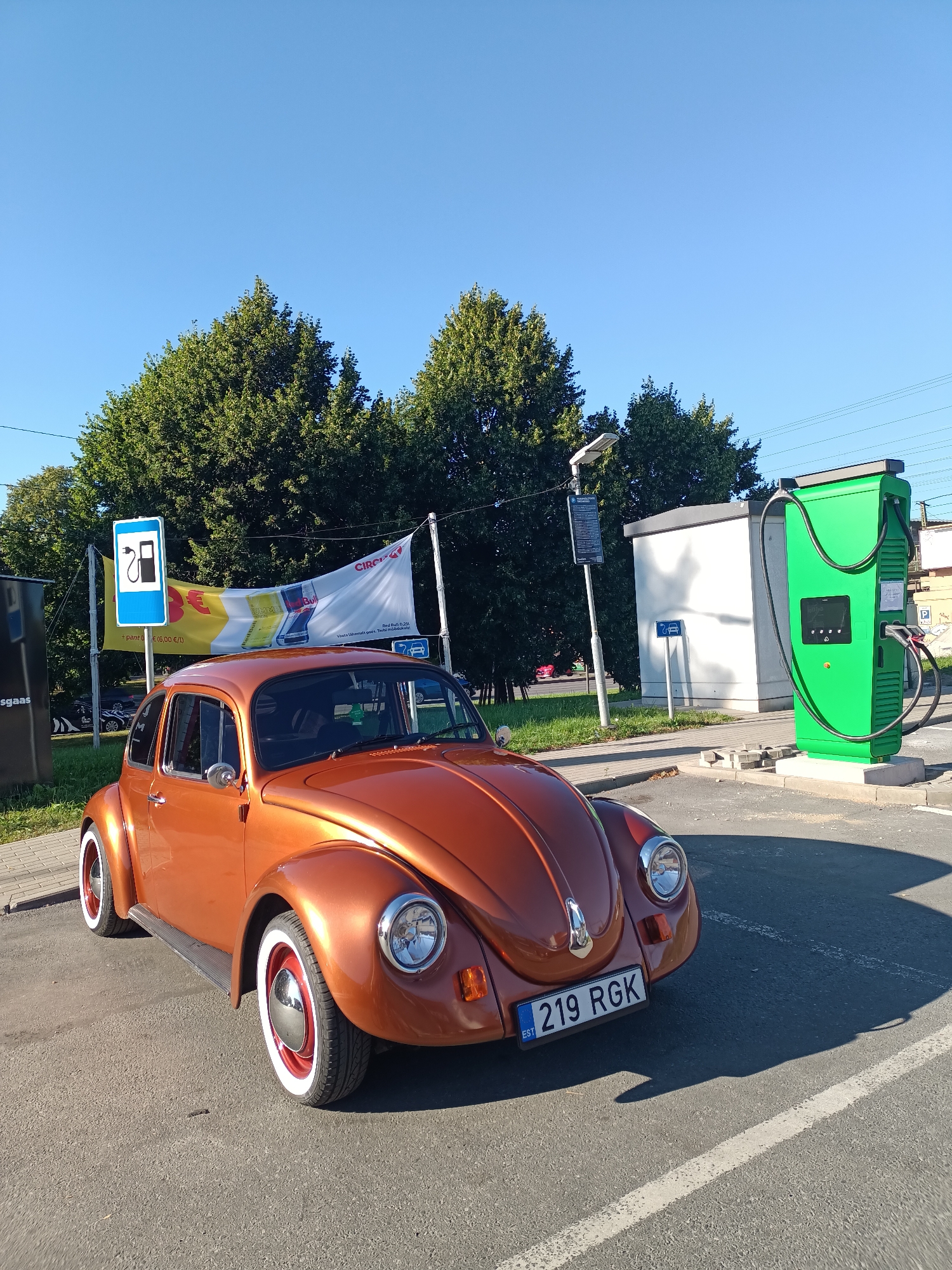 An orange Volkswagen Beetle is parked in front of a bright green electric vehicle charging station. The car, with its distinctive round headlights and chrome accents, has whitewall tires with red rims. A license plate on the front of the car reads "EST 219 RGK".
Behind the car, a white banner hangs between two poles. The banner displays advertisements for "Circle K" and "Red Bull", with some text in Estonian and a graphic of a gas pump. To the left of the banner is a blue sign with a white gas pump icon. Further to the left, a black structure with text in white is partially visible.
The charging station is tall and green, with charging cables hanging from it. To the right of the charging station, a small white building stands. A lamppost is situated between the charging station and the building.
The scene appears to be an outdoor parking lot on a sunny day. The sky is clear and blue, indicating good weather. Lush green trees form a backdrop behind the banner and structures. The overall setting suggests a modern service area or refueling station, possibly for electric vehicles, given the prominent charging station. The presence of classic car elements like the Beetle juxtaposed with the electric charging infrastructure creates an interesting visual.