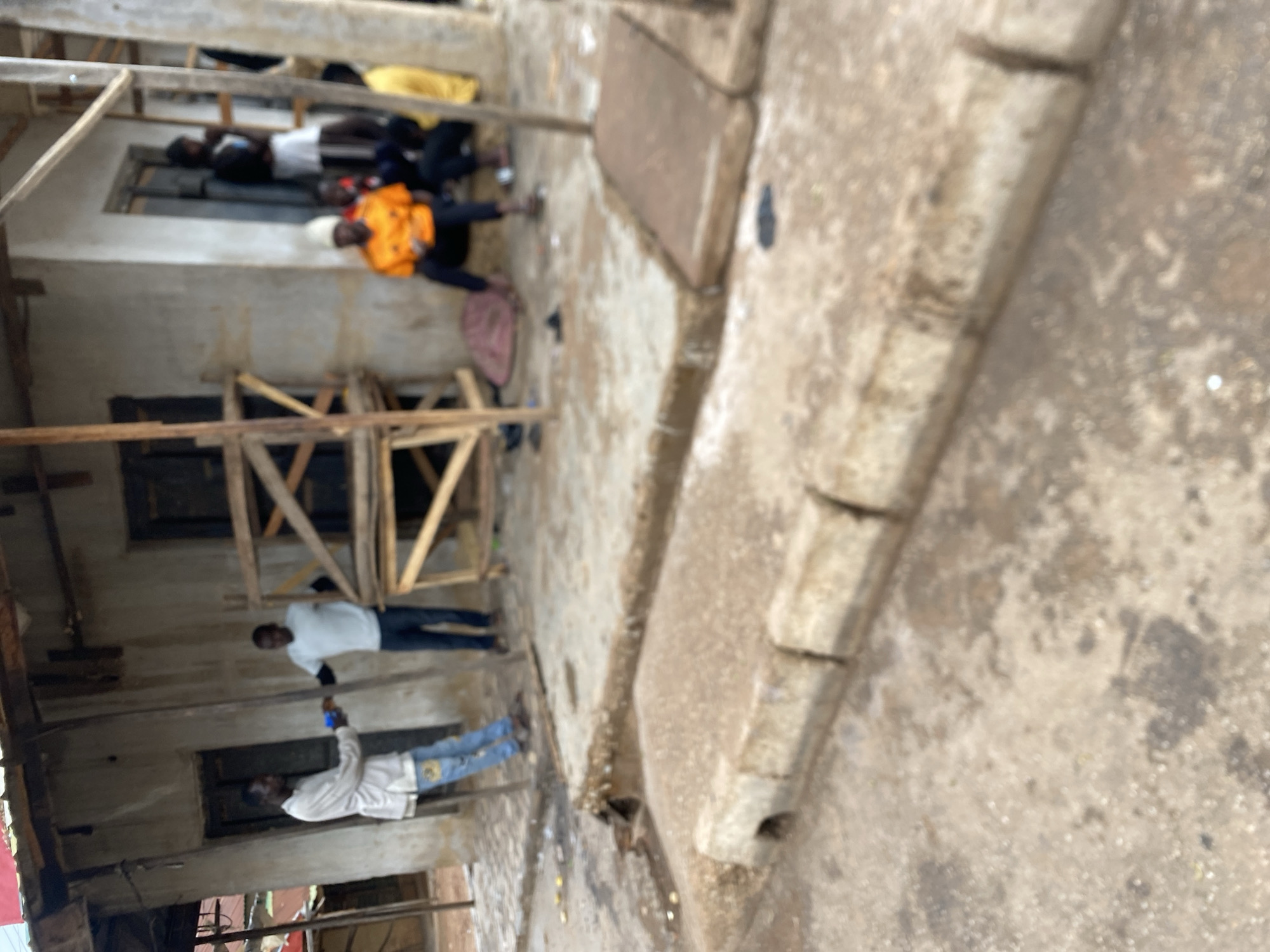 The image shows an exterior scene in Bukuru, Nigeria. Several individuals are visible, some inside a partially constructed or dilapidated building with exposed wooden beams and open window frames. There are at least seven people visible. One is wearing an orange shirt and is lying on the ground. Another person wears a white shirt with dark pants, also on the ground. The others are standing, two with light colored shirts and one of the two with light colored shirts is looking at a cellphone. The foreground features a worn concrete surface. The lighting is ambient, suggesting daytime.