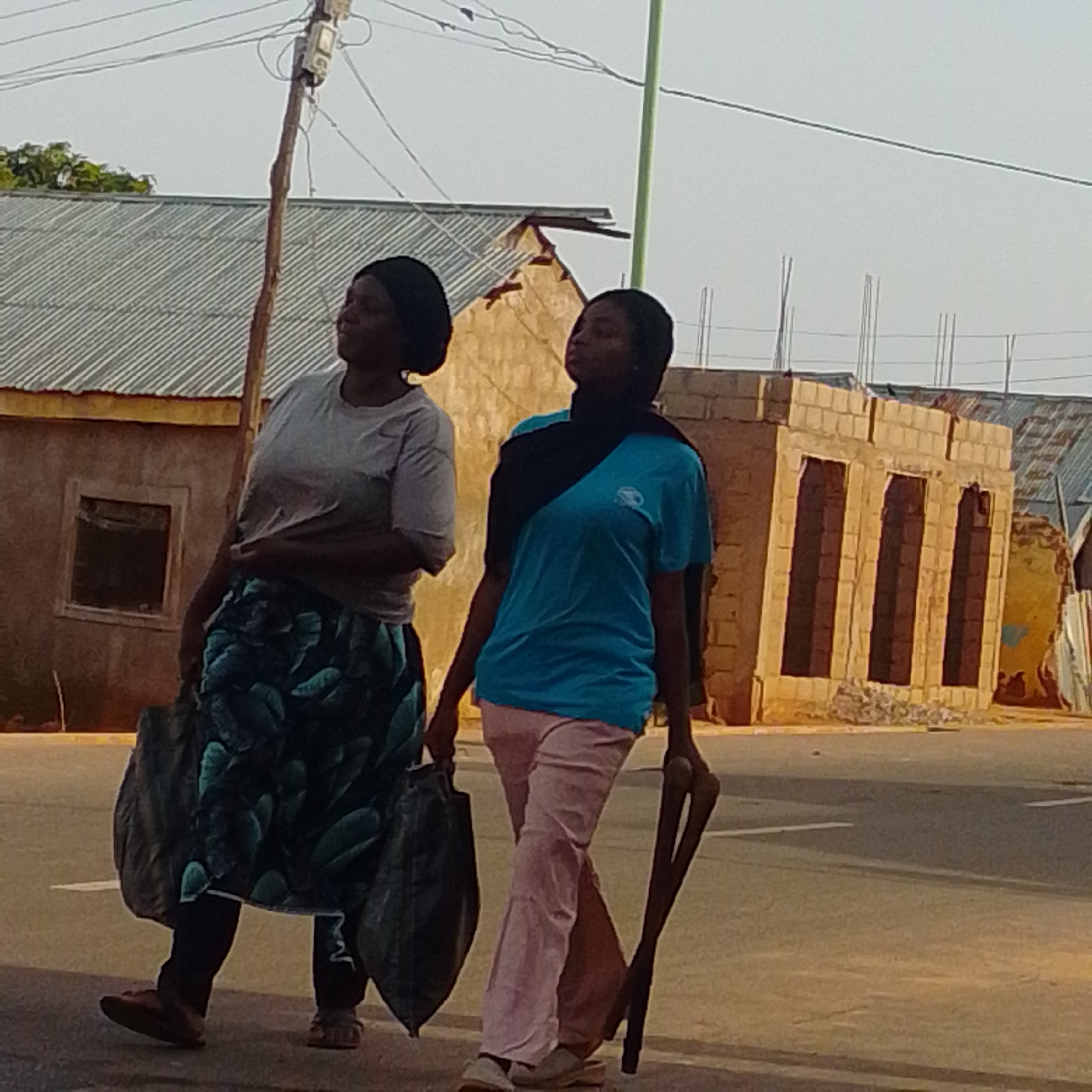 Two women are walking on a street in Tempure, Nigeria. The woman on the left wears a gray t-shirt and a patterned skirt, carrying two large black bags. The woman on the right wears a blue t-shirt and pink pants, carrying a long, dark, V-shaped object. Buildings, some under construction, are visible in the background. The sky is clear and bright.