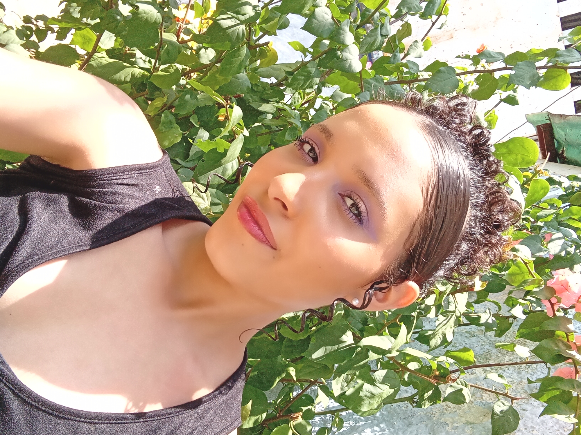 The image presents a vibrant, close-up portrait of a young woman, captured outdoors in bright sunlight. She is positioned with her head tilted, looking directly at the camera with a gentle, slight smile. Her skin appears warm-toned, and her dark, curly hair is pulled back, with some strands appearing sleek and wet, and small ringlets framing her ears. She wears a minimal pearl stud earring in her visible right ear. Her makeup includes striking purple eyeshadow, well-defined eyelashes with mascara, and glossy reddish-pink lipstick. She is dressed in a black top or tank top, visible around her shoulders and chest.
The background is dominated by lush, vivid green foliage, suggesting dense plant growth such as a bush or vine. Interspersed among the green leaves, particularly on the right side of the frame, are clusters of small pink flowers, possibly bougainvillea, indicating a warm climate. Behind the greenery, parts of a light-colored wall or structure, likely concrete or stucco, are visible, along with a glimpse of a clear, light blue sky. A dark green pot or container is also faintly visible in the upper right background. The strong, direct sunlight creates bright highlights on her face and the foliage, suggesting it is daytime with clear weather. The setting points to an outdoor area, possibly a garden or patio, consistent with the location context of Parroquia Ocumare, Venezuela. No visible text is present in the image.