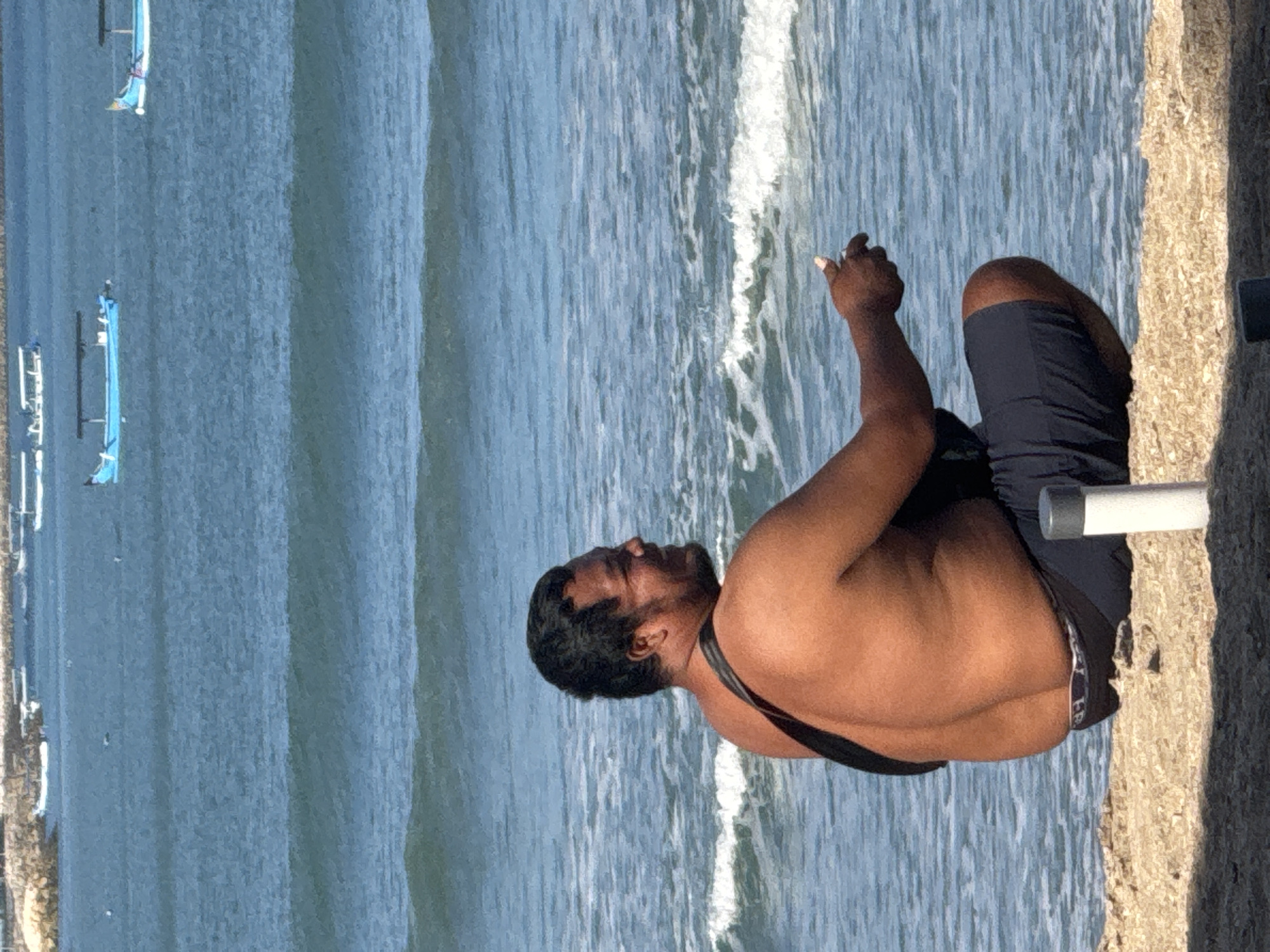 A shirtless, dark-haired man with a tan complexion is crouching on a sandy beach, facing to the right. He is wearing dark blue shorts with a patterned waistband. A black strap is draped over his left shoulder and across his back. His right hand is extended forward, and he appears to be holding something small. The ocean is behind him with gentle waves. In the distance, several boats are visible on the water. The scene is illuminated by bright sunlight, casting shadows on the sand in the foreground. This image was captured at Kuta Beach, Kuta, Bali, Indonesia.