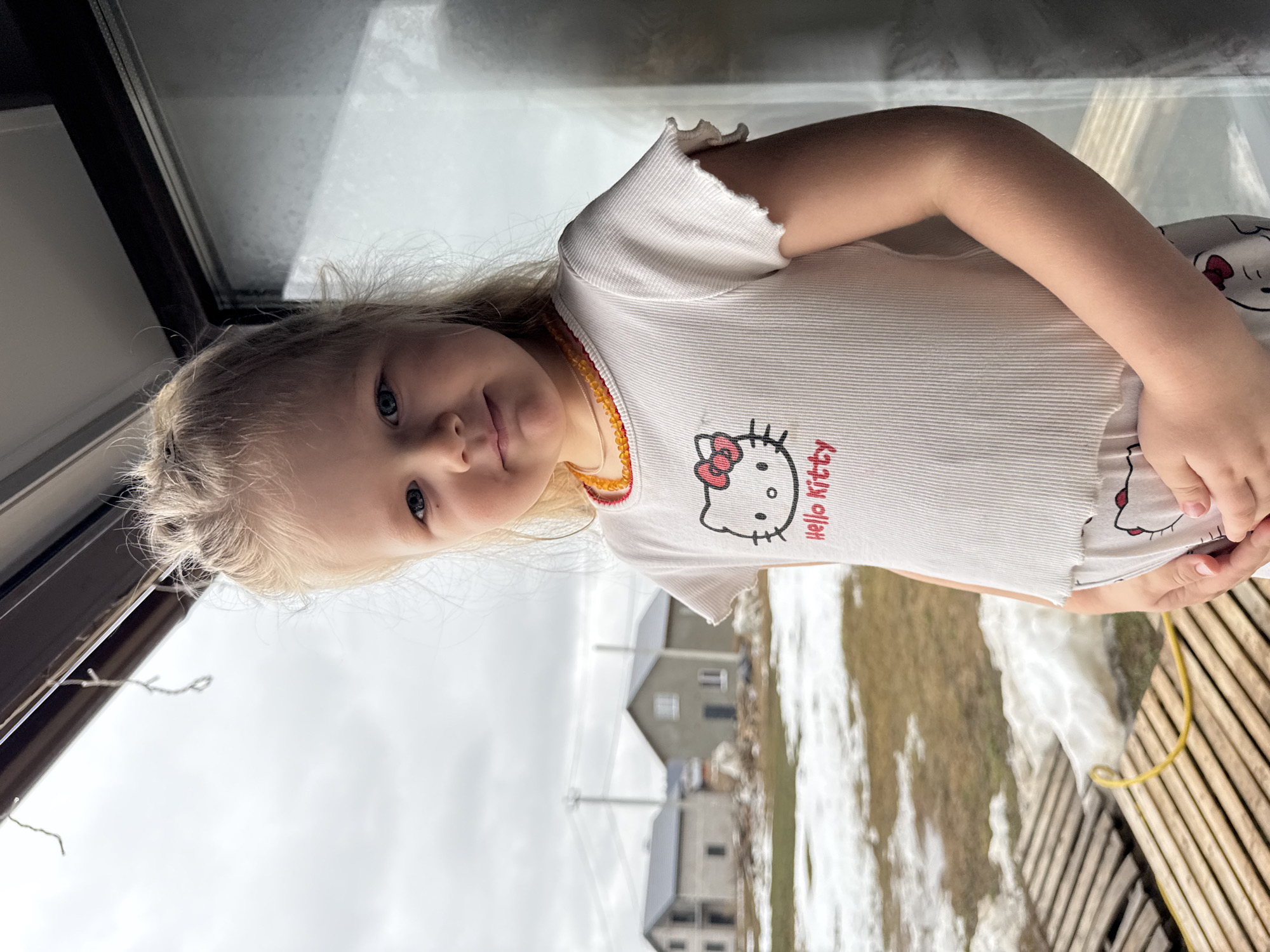 A young, light-skinned girl with blonde hair and blue eyes, wearing a white ribbed shirt with a Hello Kitty design and an orange beaded necklace, stands inside near a window. Outside the window is a view of houses and patches of snow-covered ground. The scene appears to be in Gornyi (Dvurechenskoe S/P), Russia. A wooden structure is visible near the girl. The sky is overcast.