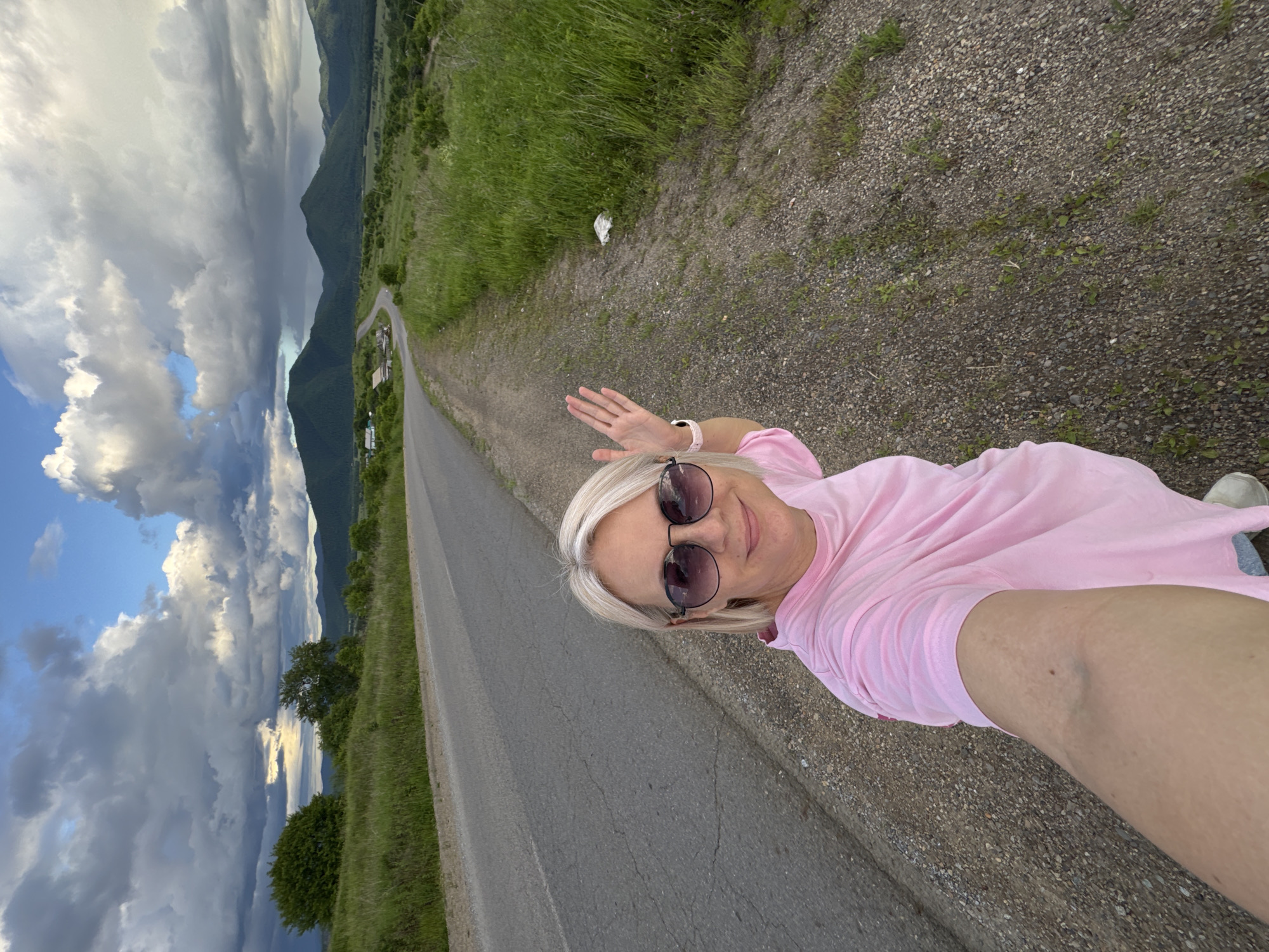 A woman with blonde hair and sunglasses takes a selfie on the side of a rural road. She is wearing a pink t-shirt. The road curves ahead, disappearing into a valley with rolling hills and mountains in the distance. The sky is partly cloudy with dramatic cloud formations. The area appears to be a natural, undeveloped landscape. There is no indication of the city of Теплоозёрск, Russia, in the image.