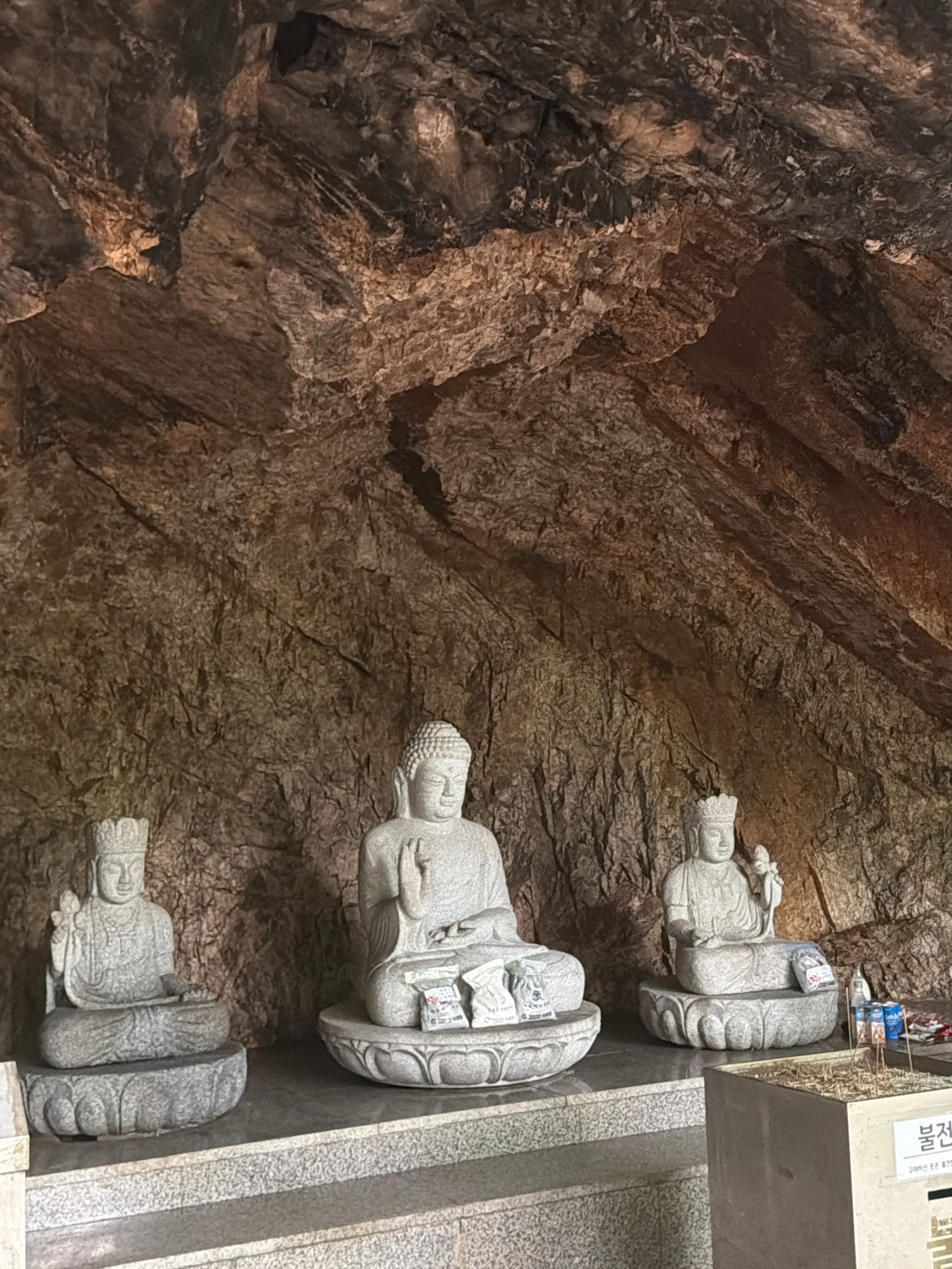 In a dimly lit cave, three stone Buddha statues are arranged on tiered platforms. The cave walls are rough and textured, with a mixture of brown and gray rock formations. The statues are seated on lotus-shaped pedestals. The central statue is the largest and has its hands in a gesture of prayer or blessing. The statue on the left is smaller and holds a lotus flower in its left hand. The statue on the right is also smaller and holds a small object in its left hand, possibly a bud. Offerings, including small bags and prayer papers, are placed on the pedestals. To the right of the statues, there is a planter filled with incense sticks and a sign with Korean writing that reads "불전" (Buljeon), which means "temple offering" or "donation". The scene appears to be a place of worship, likely a Buddhist temple or shrine, within a cave setting in Dongducheon-si, South Korea.