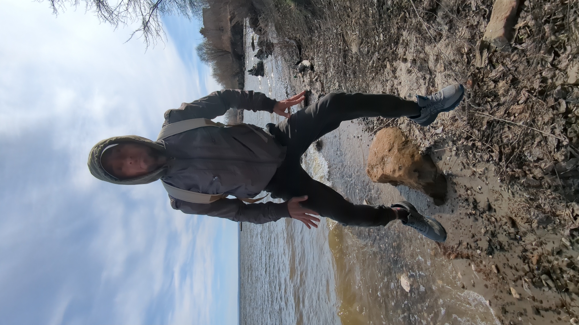A full-length, vertically oriented shot captures a person standing on a muddy, rocky riverbank or lakeside. The subject, a male figure, is dressed in a dark grey hooded jacket with the hood up, dark trousers, and grey athletic shoes. Light-colored straps, possibly from a backpack, are visible over his shoulders. He is captured in a dynamic pose, with his left leg extended forward and slightly lifted, and his arms bent at the elbows, hands open and raised to his sides, as if in motion or balancing. He looks towards the camera with his face partially obscured by the hood.
The setting is an outdoor natural environment. To the left of the person, a body of water, likely a river or large lake, flows with murky, brown-tinted water, showing small ripples and waves. The shore on the right is covered with wet soil, small rocks, and dried leaves and twigs. A larger, light brown rock sits on the immediate bank near the person's right foot. In the background, bare-branched trees line the distant bank under a partly cloudy, light blue sky. The overall atmosphere suggests a cool, possibly early spring or late autumn day. No visible text is present in the image.