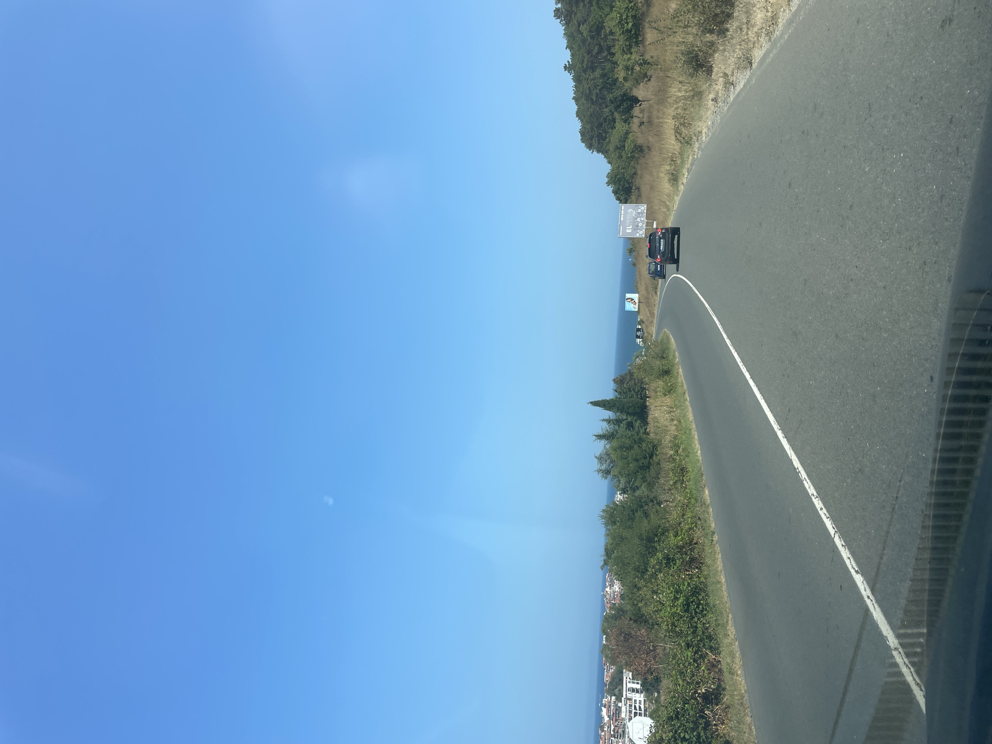 The image captures a view from a vehicle traveling on a paved road that curves to the left. The road has a white line marking the lane. On the left side of the road, there is a town with white buildings and some greenery, followed by the sea. On the right side of the road, there are dry, grassy hills dotted with trees. The sky is clear and blue. In the distance, two cars are visible on the road, with one black SUV ahead and a dark car behind it. There are also billboards visible along the right side of the road. The scene appears to be a coastal road near Sozopol, Bulgaria, on a sunny day.