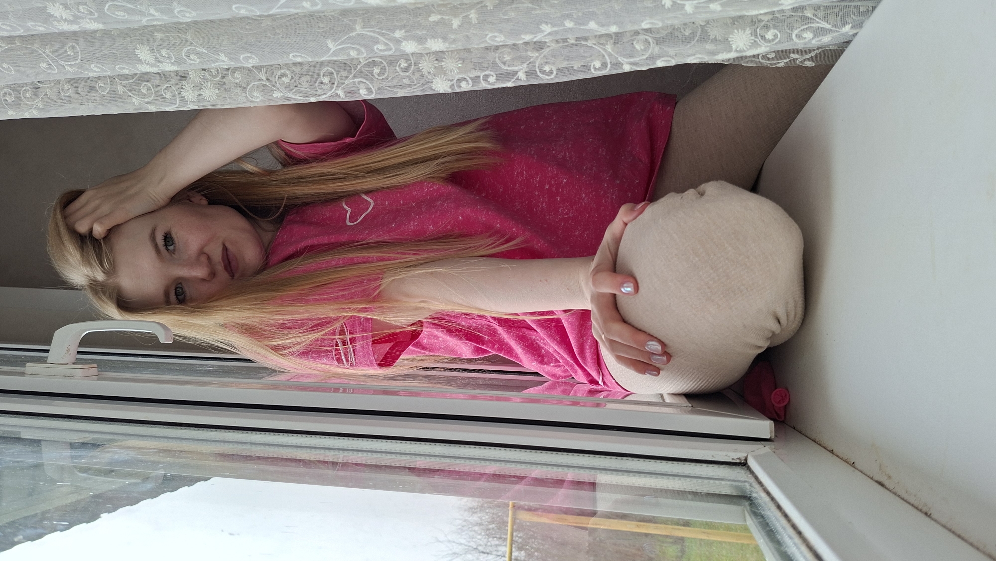 A woman with blonde hair is lying on a windowsill with a stuffed animal. She is wearing a pink shirt and light-colored leggings. The window is open and there is a view of trees and grass outside. A white curtain hangs above her. The location is in the city of Большой Букор, Russia.