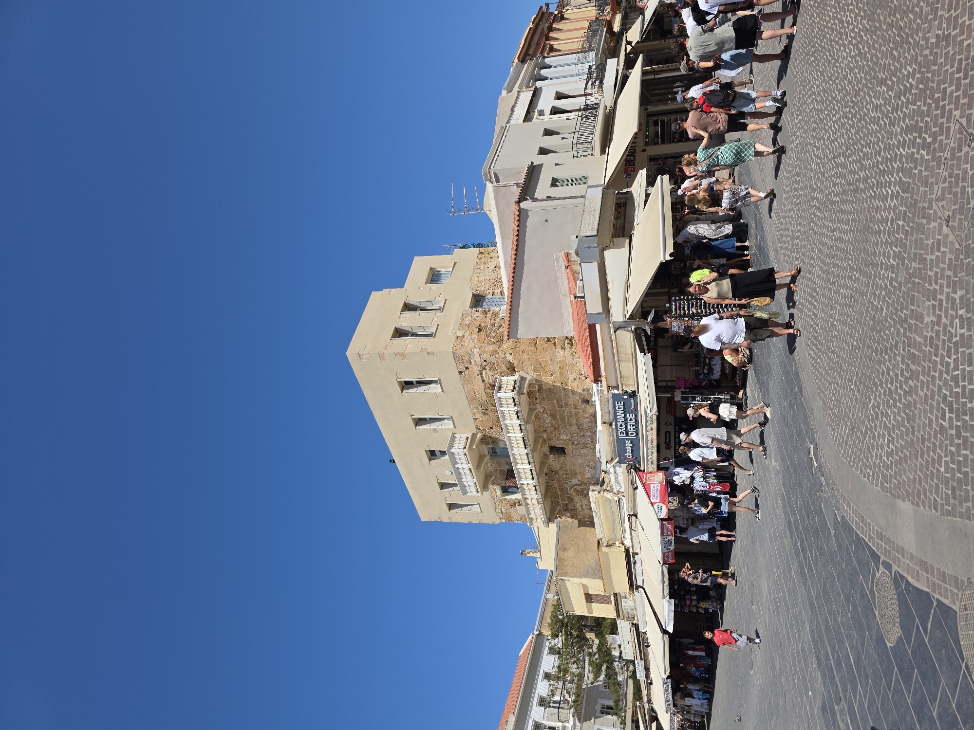 The image depicts a bustling street scene in the city of Chania, Greece, likely within the Venetian Harbor area, characterized by its historic architecture and cobblestone pavements. The primary focus is on a tall, multi-story building constructed with light-colored stone at its base and plastered upper floors, featuring white-railed balconies. In front of this structure, a row of shops with beige awnings extends along the street. The shops display various goods, and some signage is visible, including "EXCHANGE OFFICE" and "BEAUTY".
The street is paved with a combination of dark gray square tiles and reddish-brown cobblestones. Numerous people are walking along the street, some browsing the shops, others moving through the square. They are dressed in casual summer clothing, suggesting warm weather. A young boy in a red shirt and gray shorts is walking alone in the foreground.
The sky is a clear, bright blue, indicating a sunny day. The overall atmosphere is one of lively commercial activity within a historic Mediterranean setting.