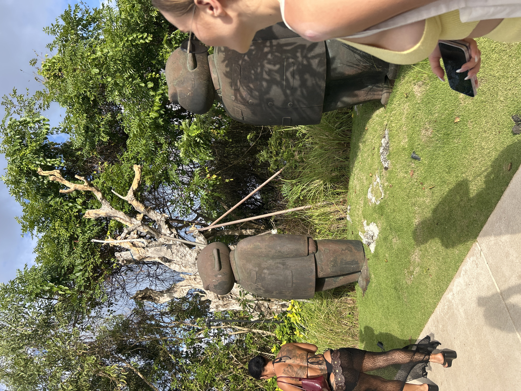 In Kuta Selatan, Indonesia, two large, dark-colored statues resembling stylized figures in formal wear stand on a grassy lawn. The statue on the left is viewed from the side, and the statue on the right is viewed from the back with its head turned to the right. A woman with dark hair and tattoos on her back, wearing a black bikini top and a sheer black skirt, walks away from the statues on a concrete path. Another woman, whose right side is in the foreground, looks up and to the right, holding a phone in her right hand. She is wearing a yellow top. Lush green trees and vegetation surround the statues and the women. The scene is illuminated by daylight, casting shadows on the grass and the path.