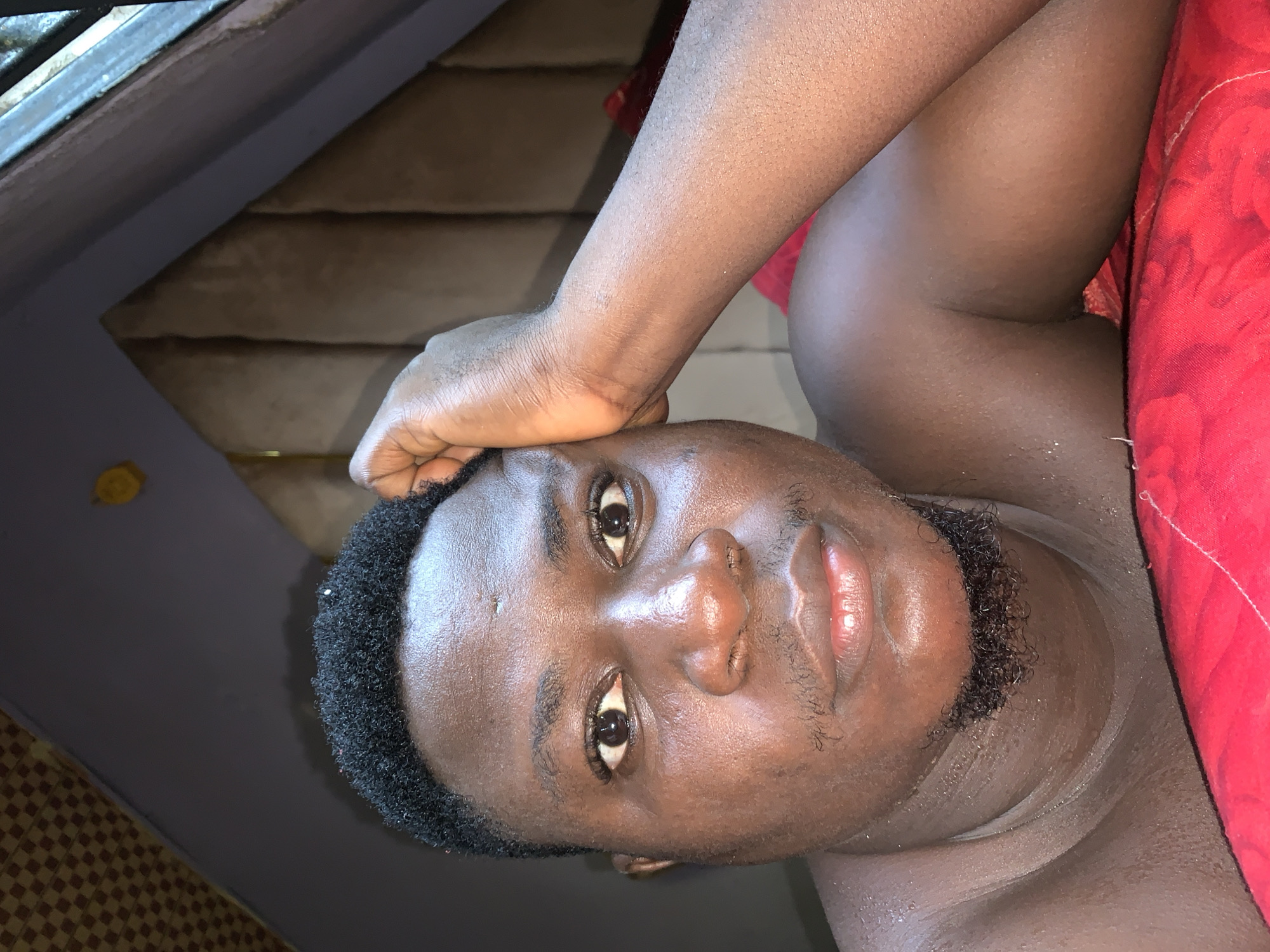 This close-up portrait features a dark-skinned individual, likely male, lying down indoors in Akwanga, Nigeria. The person is positioned with their head slightly tilted, looking directly at the camera with an open, direct gaze. Their left arm is bent, and the hand rests on their forehead, adding to a casual, relaxed pose.
The individual has short, dark, curly hair and a trimmed goatee or short beard. Their skin exhibits a noticeable sheen, possibly from sweat, oil, or specific lighting. Their eyes are dark and prominent.
To the right of the frame, a vibrant red fabric, possibly a blanket or part of an outfit, is visible, partially covering the person's upper body. The background elements suggest an informal indoor setting. In the upper left, a dark, angular structure, potentially part of a window frame or interior architectural detail, is visible. Behind the person's head, a textured surface made of horizontal wooden planks or panels adds depth to the scene. Towards the bottom left, a portion of a tiled or checkered floor can be seen, along with a small, circular yellow object.
The lighting is soft and even, highlighting the subject's features without strong shadows, consistent with indoor ambient light. There is no visible text in the image. The overall impression is a personal and direct portrait.