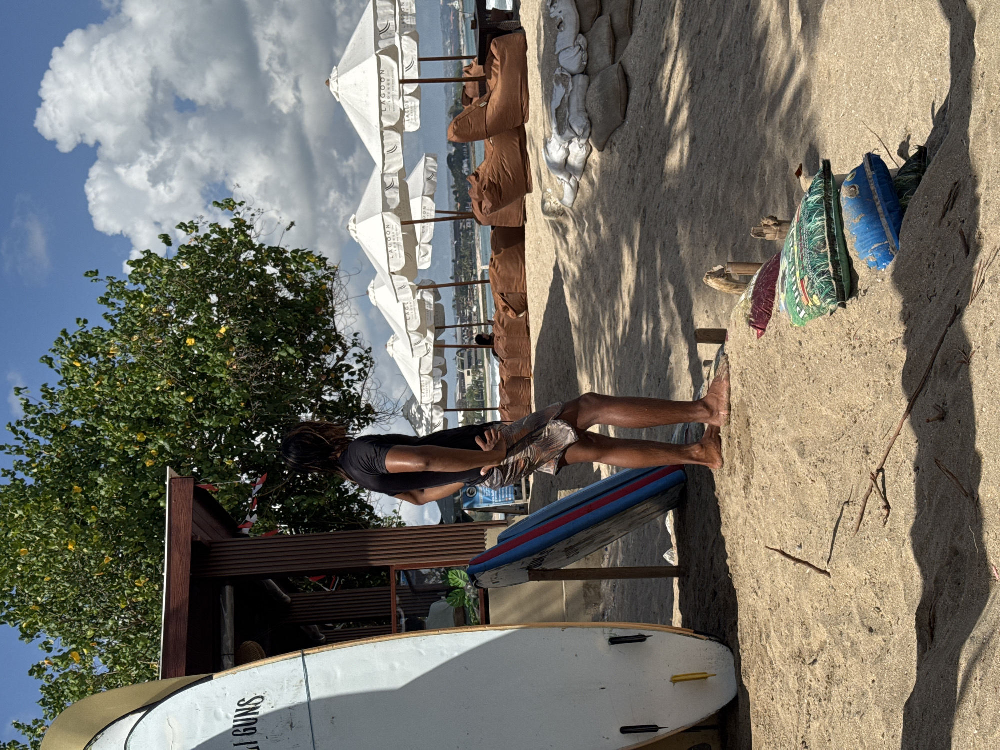 A lone man stands on a sandy beach in Kuta, Indonesia, looking out towards the ocean. He is wearing a black t-shirt and patterned shorts, with his hands on his hips. In front of him, several surfboards are propped up. Behind the man, there are several white umbrellas providing shade over brown beanbag chairs, suggesting a beach lounge area. The beach is bordered by trees and the ocean can be seen in the distance under a partly cloudy sky. The scene conveys a sense of relaxation and leisure, typical of a popular beach destination.