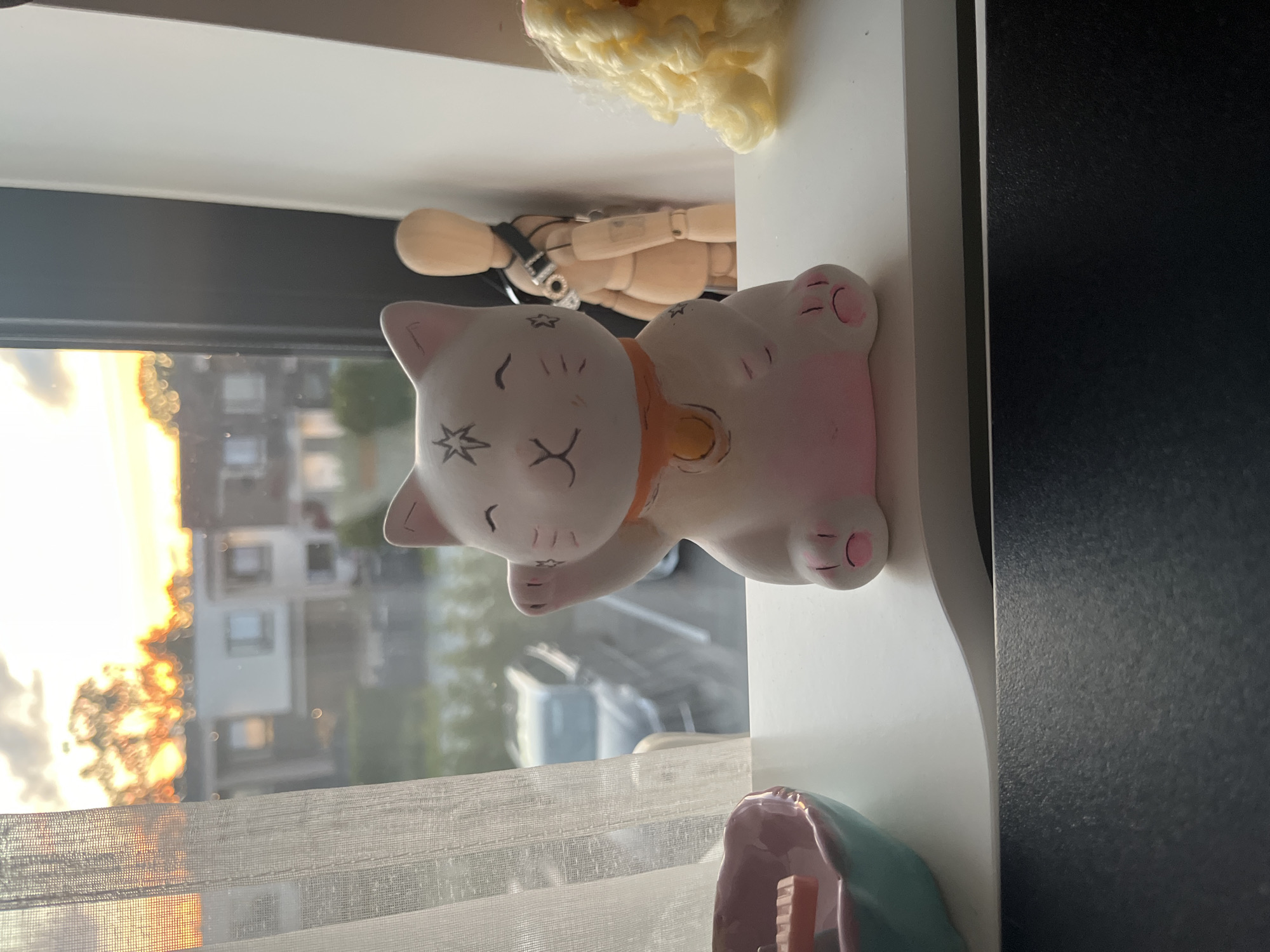 A maneki-neko figurine, a white cat with pink accents and an orange collar, sits on a white ledge overlooking a city view at sunset. The background shows buildings and trees illuminated by the setting sun. To the left of the maneki-neko, a small pink and blue dish with an orange object is visible. Behind the figurine, a wooden mannequin is positioned with a strap across its chest. To the right, a fluffy yellow object is partially in frame. The scene appears to be indoors, with a sheer curtain to the left. The maneki-neko is the primary subject, positioned centrally on the ledge.