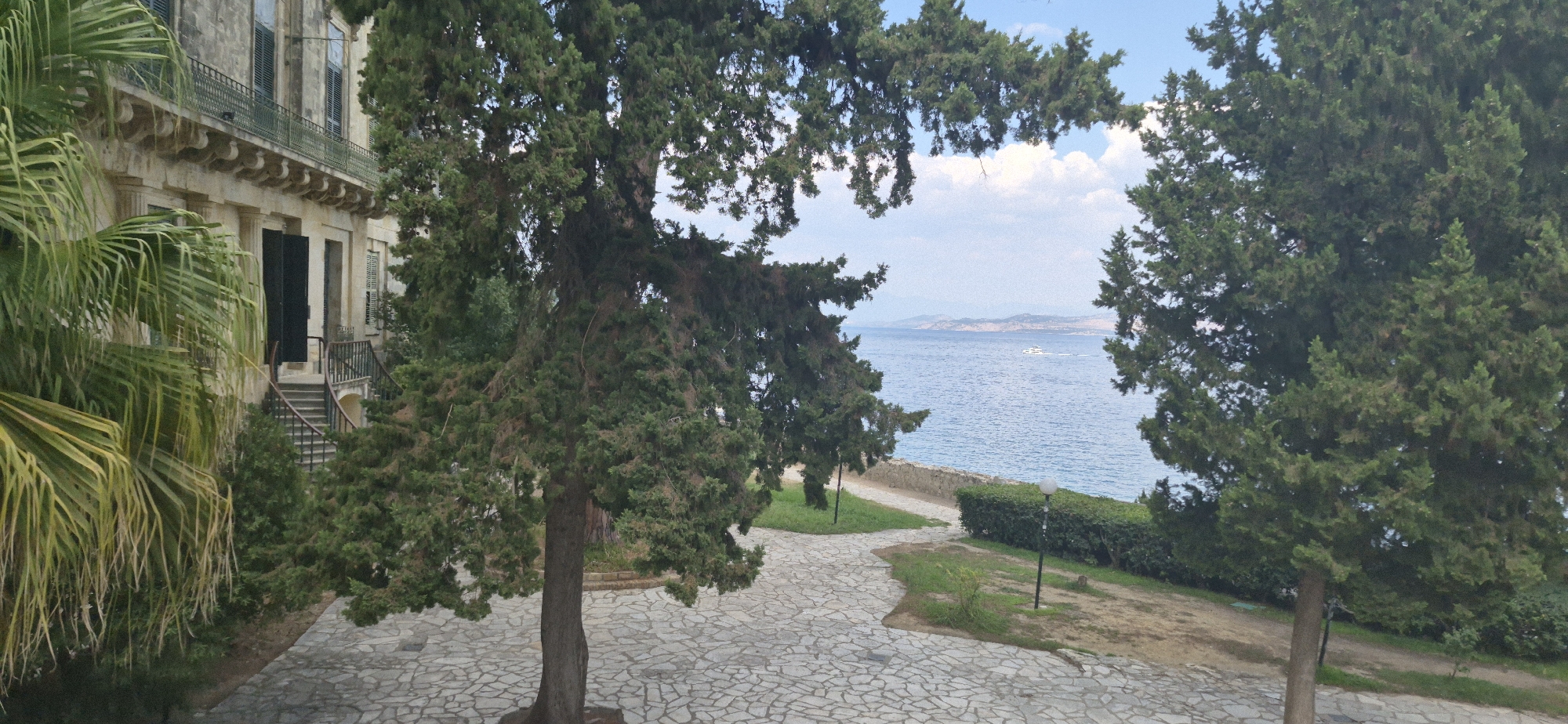This image depicts a scene in the city of Corfu, Greece, likely in the vicinity of the Old Fortress or a similar historical estate overlooking the sea. In the foreground, a paved pathway made of irregularly shaped stones winds through a garden area. Several large evergreen trees, possibly cypresses or pines, dominate the midground, their dense foliage partially obscuring the view of the sparkling blue sea and a distant coastline under a partly cloudy sky. To the left, a section of a grand, neoclassical building with stone columns, arched windows, and a balcony with an ornate railing is visible. A staircase leads up to the building's entrance. On the right side of the image, a low stone wall separates the garden from the sea, and a neatly trimmed hedge runs along its edge. A small white boat is visible on the water in the distance. The overall atmosphere is serene and picturesque, suggesting a historical and well-maintained garden setting.