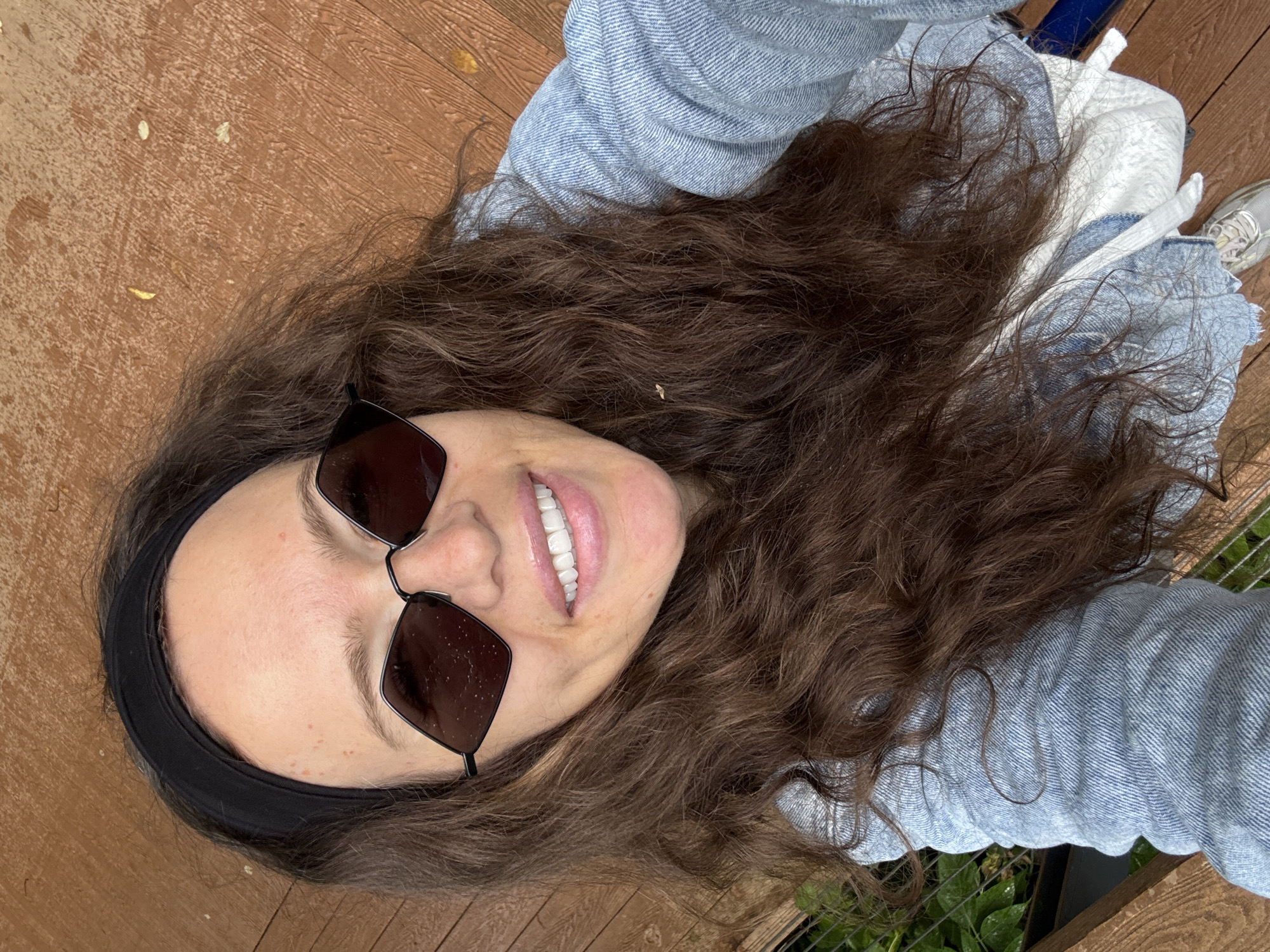 A woman with long, dark, curly hair and a black headband is looking at the camera. She is wearing diamond-shaped sunglasses and a light blue denim jacket over a white garment. Her teeth are visible as she smiles. The background appears to be a wooden deck with some leaves scattered on it, and a glimpse of greenery and a fence to the left. This scene is not in Moscow, Russia.
