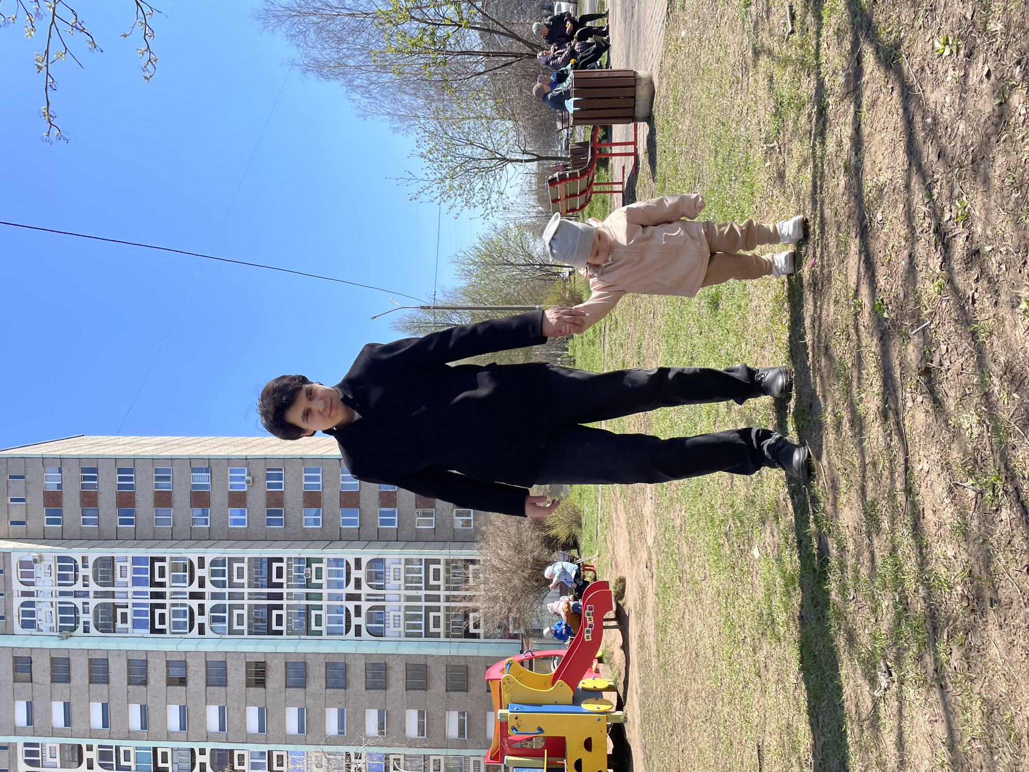 A man and a child are holding hands in a park-like setting, potentially in Izhevsk, Russia. The man is wearing a black suit and black shoes, standing with the playground equipment in the foreground. The child, dressed in light-colored clothing and a grey hat, is slightly ahead, pulled forward by the man. In the background, there is a large residential building and people sitting on benches. The ground is a mix of grass and dirt, suggesting a well-used public space. The sky is clear and blue.