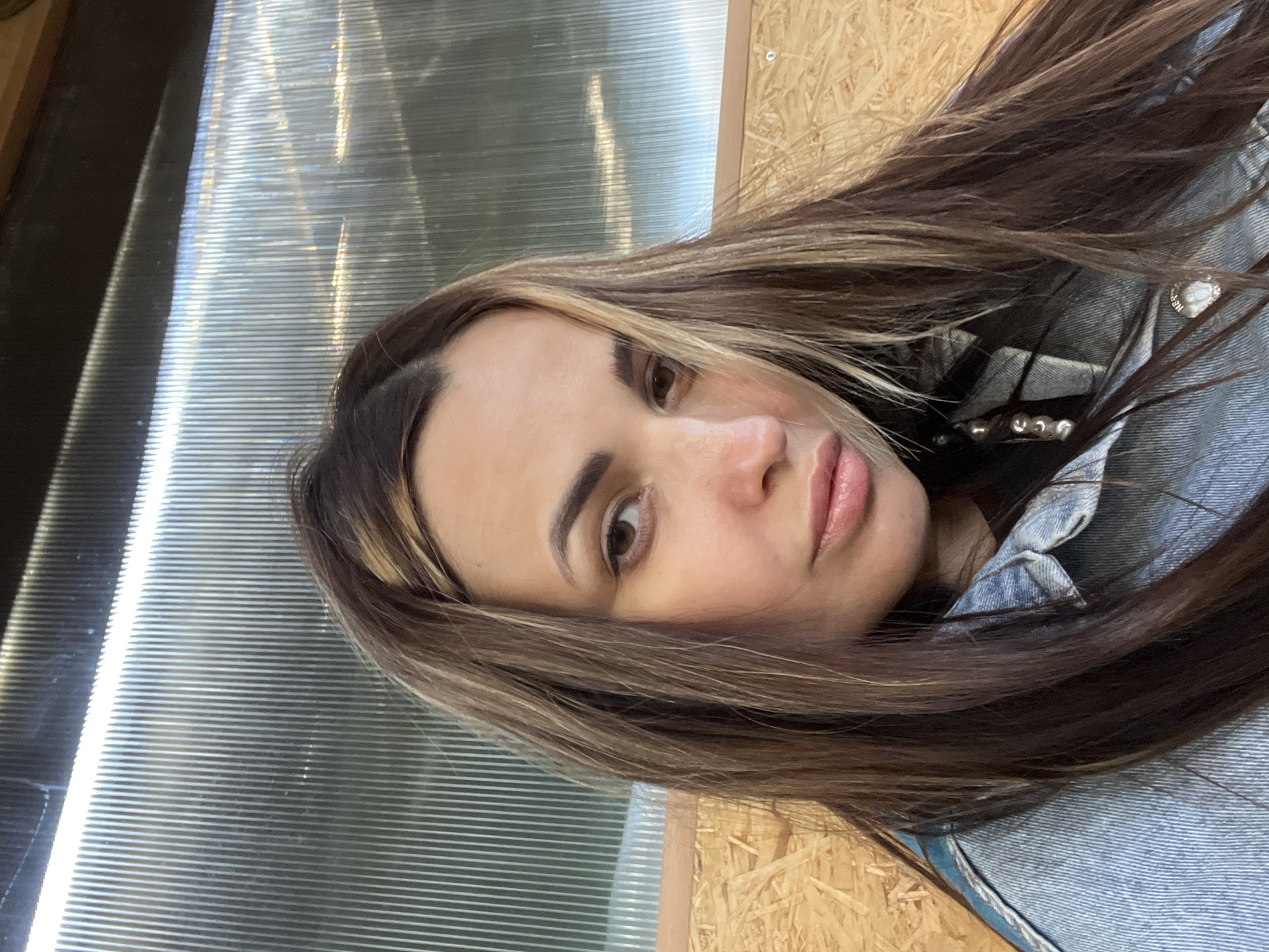 A woman with long, brown hair with blonde highlights is looking directly at the camera. She is wearing a denim jacket and has fair skin. The background appears to be a combination of corrugated plastic and plywood. The location is unknown.