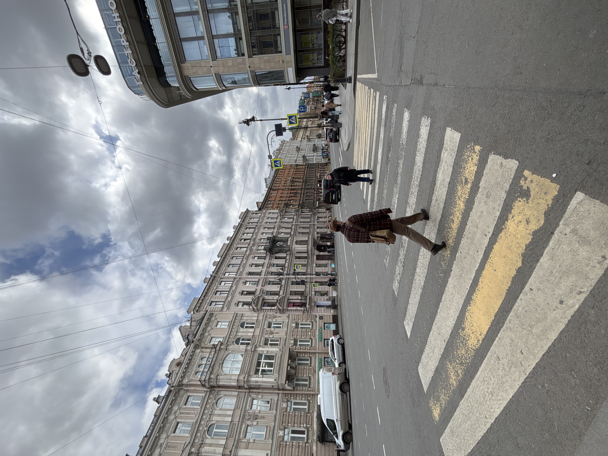 The image captures a street scene in Saint Petersburg, Russia, featuring pedestrians crossing a zebra-striped crosswalk. The pavement of the crosswalk has yellow accent lines. The buildings surrounding the street have multi-story facades with ornate architectural details. The sky is overcast with thick, textured clouds. Several streetlights are visible along with power lines running overhead. A white van and a car are parked along the street. People are interacting with the street environment, crossing the road on foot.