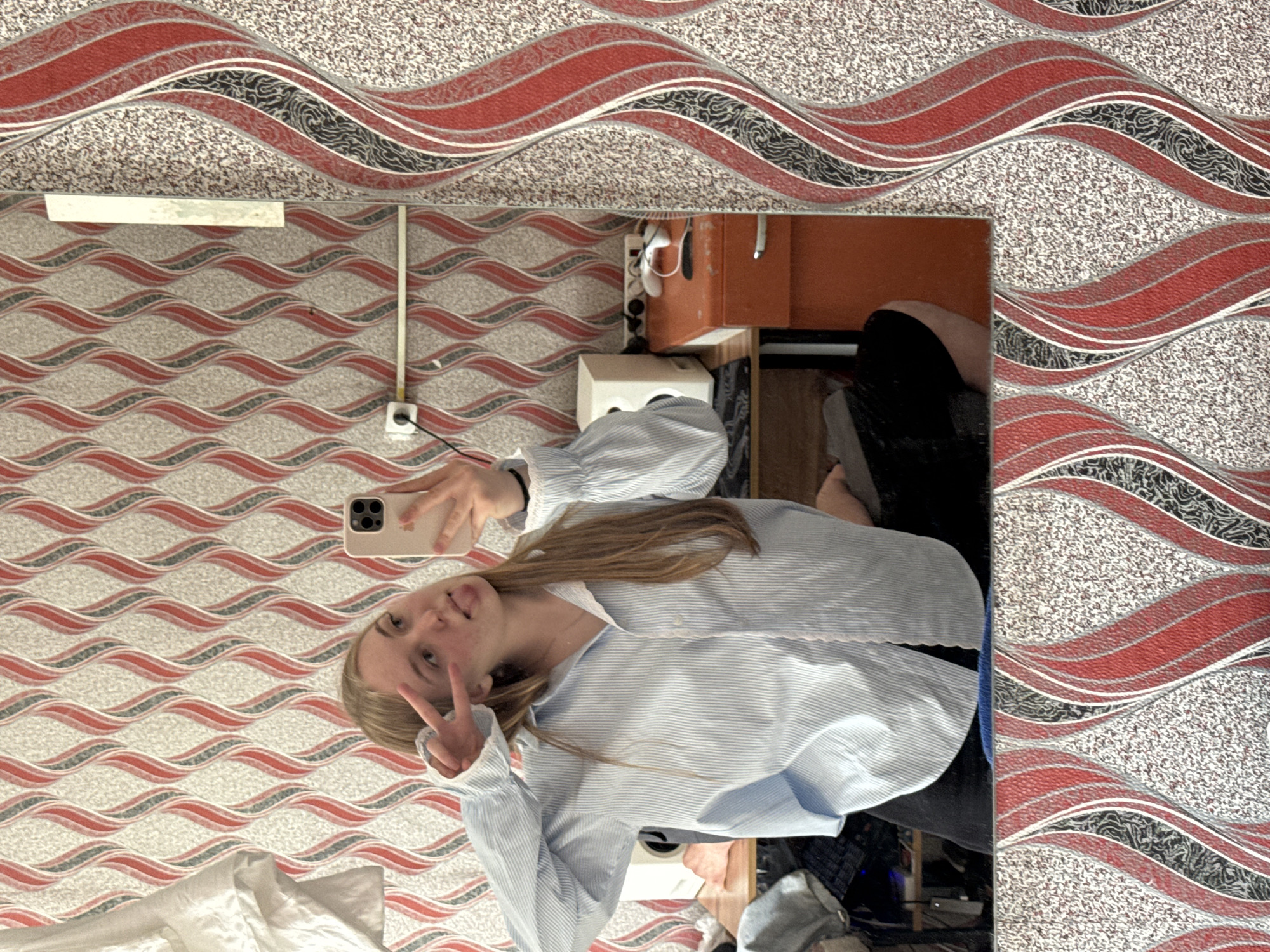 A young woman takes a selfie in a mirror. She has light-colored hair and is wearing a blue and white striped shirt. She is holding up two fingers in a peace sign. The background is a patterned wall with red, gray, and white waves, indicating the image was taken in a domestic setting, likely in Perm, Russia based on environmental cues and the wallpaper style often seen in Russian homes. The room includes a cabinet, and other indiscernible objects can be seen in the mirror's reflection.