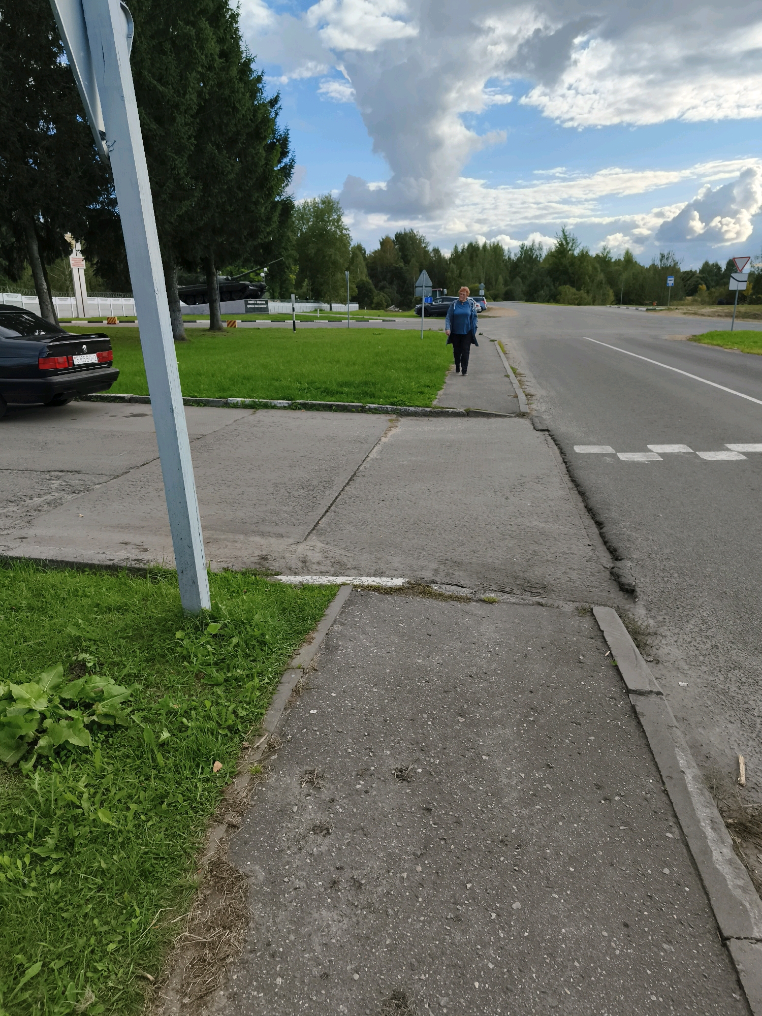 A woman with auburn hair is walking on a concrete sidewalk adjacent to a paved road. She is wearing a blue jacket and dark pants. The sidewalk is cracked and uneven. To her left is a patch of green grass and a white pole. To her right, the road has white lane markings. In the distance, there is a line of trees and a blue sky with white clouds. A black car is parked on the grass to the left of the sidewalk. There are several traffic signs visible in the background. The scene appears to be in a semi-rural area, possibly on the outskirts of a town. The location is identified as Горскі сельскі Савет, Belarus.