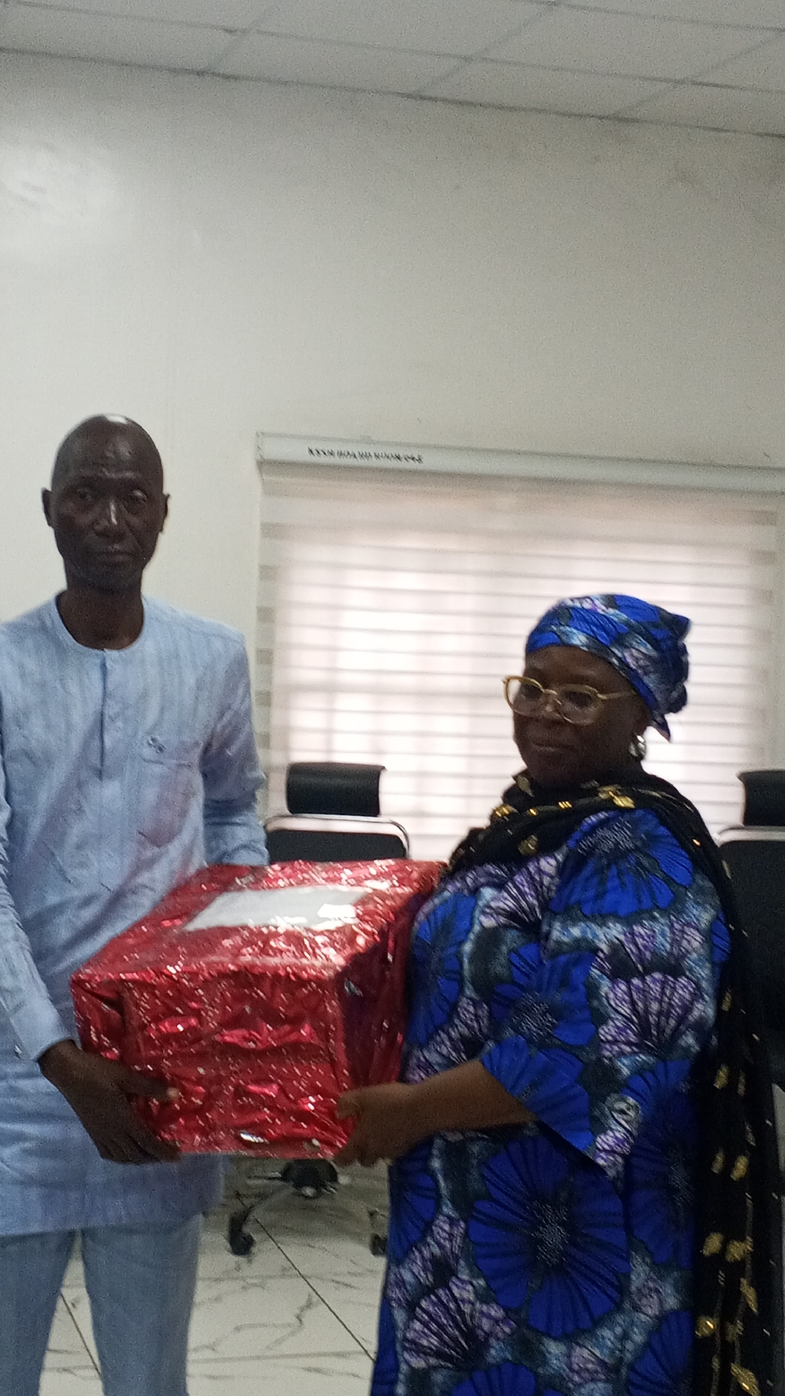 The image features two individuals, a man and a woman, standing side-by-side in what appears to be an office or conference room, jointly holding a large gift package.
The man on the left is dark-skinned and bald, wearing a light blue, long-sleeved traditional Nigerian tunic with a buttoned placket. He has a neutral expression and looks directly at the camera.
The woman on the right is also dark-skinned, wearing glasses and a blue and purple patterned dress or top, complemented by a matching blue patterned headwrap (gele). A dark shawl with gold patterns is draped over her left shoulder. She has a subtle smile and is looking slightly away from the camera.
Both individuals are presenting or receiving a large rectangular gift wrapped in shiny red paper, which they hold together. The setting is an indoor space with light-colored walls and a tiled floor featuring a light grey and white pattern. In the background, dark office chairs and a table are partially visible. White horizontal window blinds can be seen behind the woman. The ceiling is made of white acoustic tiles. Faint, blurry text, resembling "XXXX XXXXXXX XXXXXX XXXX" or similar capitalized letters, is visible on or above the blinds but is not decipherable.
The scene suggests a formal or semi-formal event, possibly a gift exchange, award presentation, or farewell, taking place in Lokoja, Nigeria. The attire of both individuals indicates a cultural or traditional context.