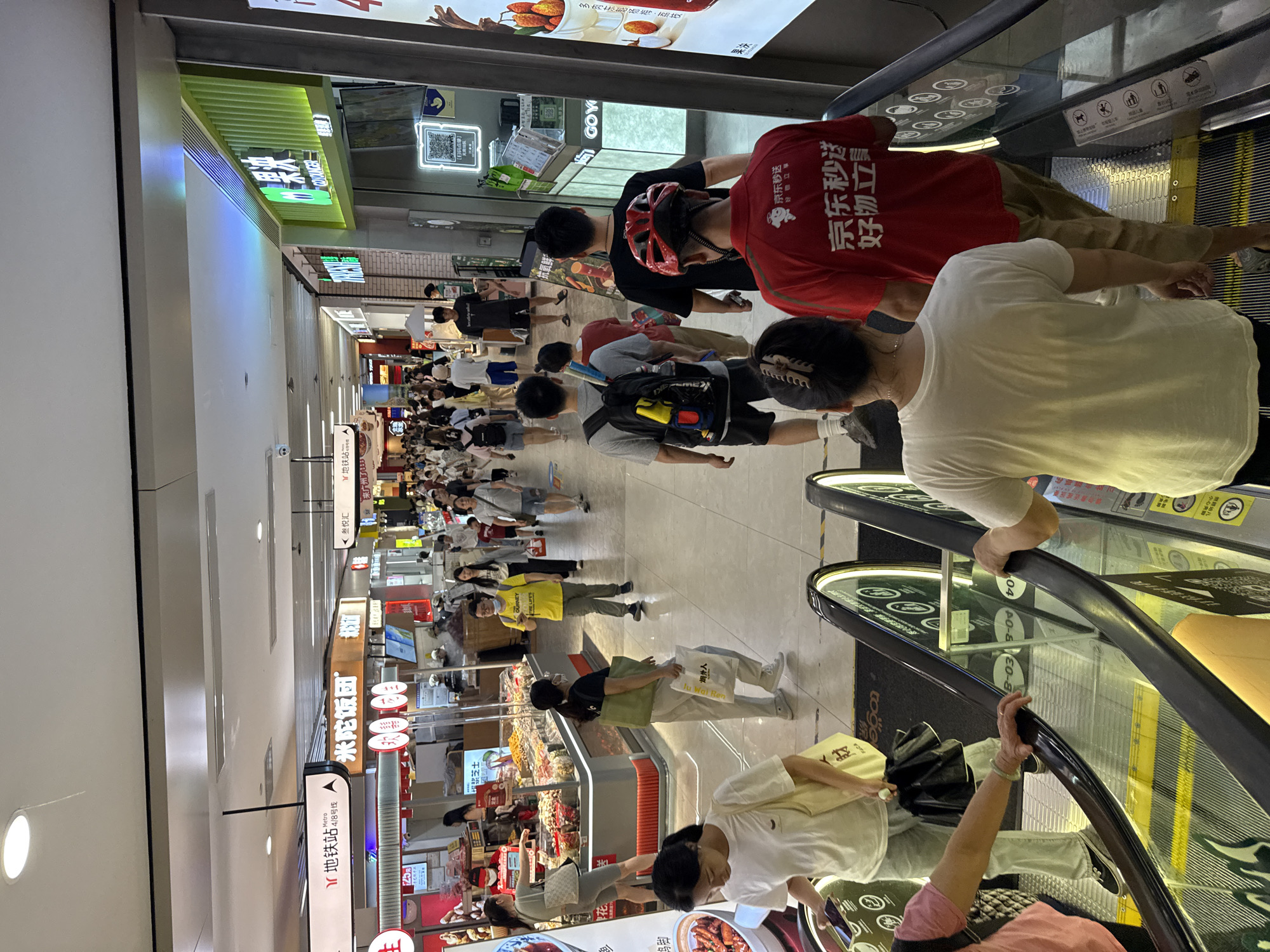 The image depicts an indoor scene, likely a shopping mall or a transit hub, with a focus on an escalator. Several people are visible, most of them walking or standing. In the foreground, two individuals are on an escalator, with their backs to the camera. One person on the right is wearing a red shirt with Chinese characters that translate to "JD Express, Good Things Stand." The other person on the left is wearing a white shirt and has a shopping bag. Behind them, more people are walking along a wide corridor, some browsing stalls or shops lining the walkway. The environment appears to be brightly lit, with signs and advertisements visible in the background. Some of the signage includes Chinese characters, suggesting a location in China. Specifically, the sign "地铁站 4/8号线" (Metro Station Lines 4/8) indicates a connection to the metro system. Another sign in green with "果" (fruit) in white text is also visible. The overall impression is of a busy commercial area. The location is identified as Haizhu District, China.