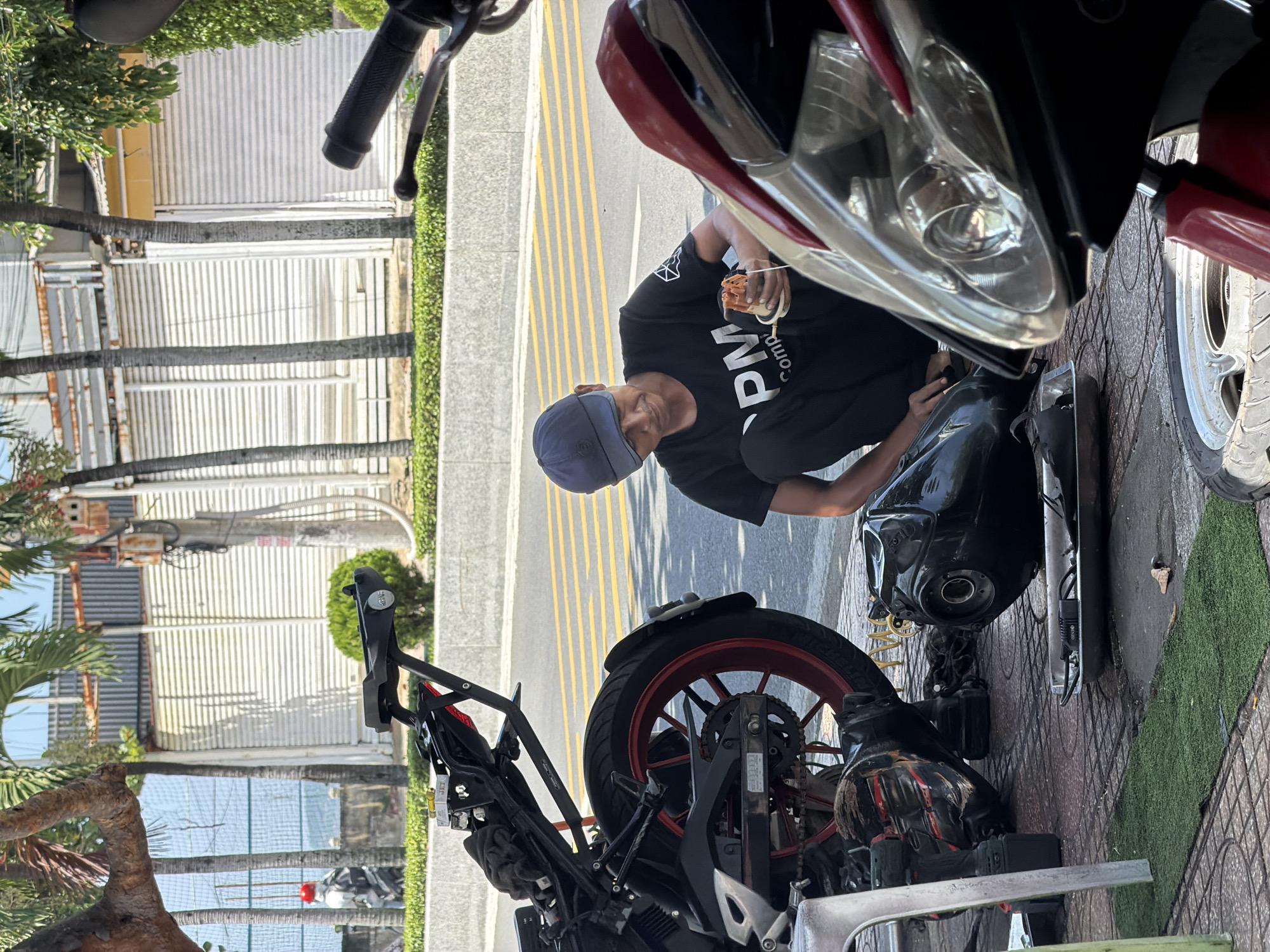 A man in a black t-shirt and a blue baseball cap is kneeling on a tiled sidewalk in front of a disassembled motorcycle. He appears to be working on a part of the motorcycle, holding a small, orange object in his hands. The motorcycle's fuel tank is lying on the ground next to him, and other parts are scattered around. Tools, including a drill, are also visible. The scene is outdoors, with palm trees and buildings in the background, suggesting an urban environment, possibly near the coast in Nha Trang, Vietnam. The ground is paved with tiles and a strip of artificial grass. The overall impression is of a roadside repair or maintenance operation.