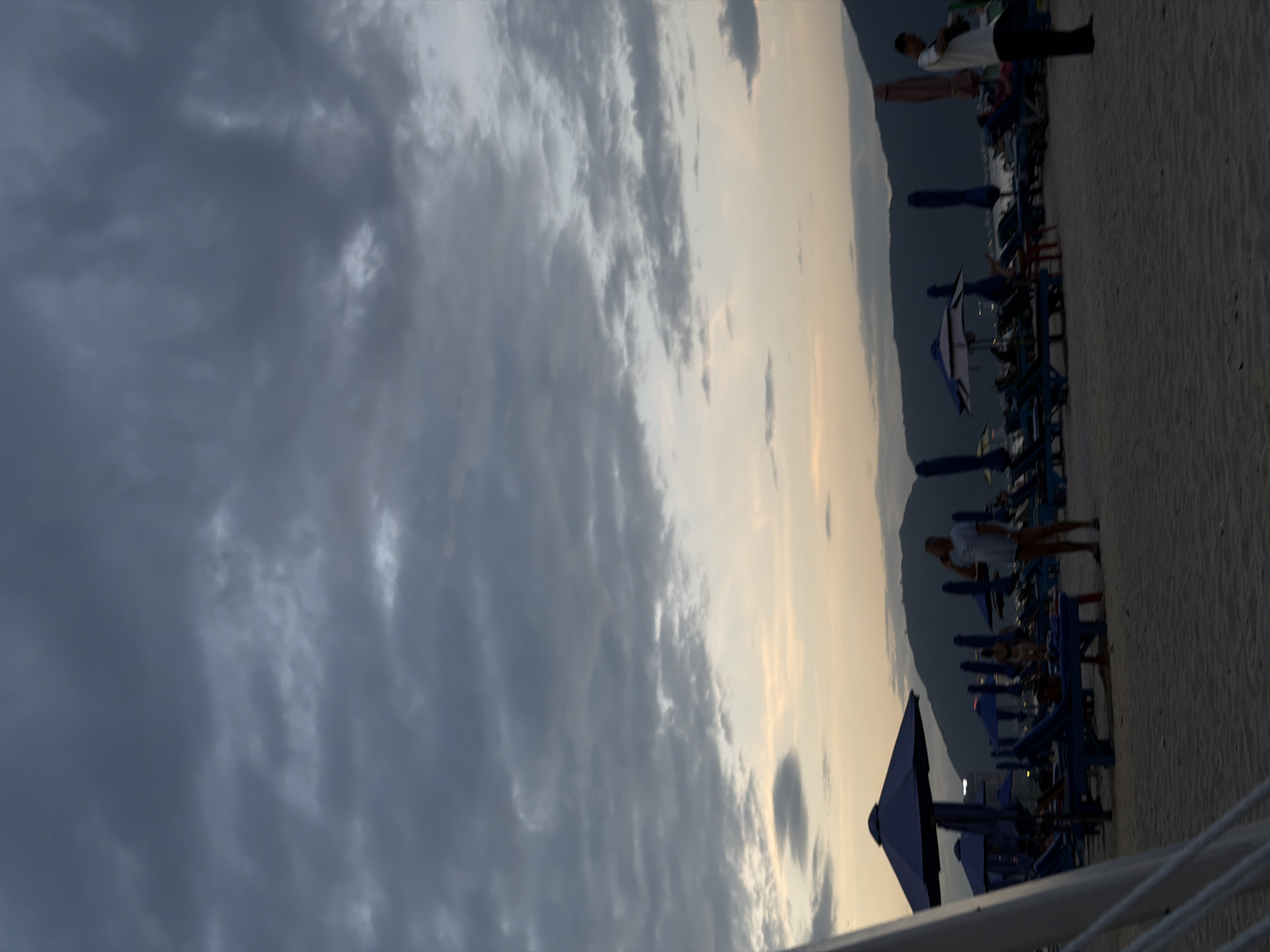 This is a medium-shot, vertical, eye-level photograph of a beach in Da Nang, Vietnam, during dusk. The sky is filled with dark, overcast clouds, transitioning to a lighter, pale orange hue near the horizon. The sea is visible in the background, as are dark, silhouetted mountains. In the foreground, a sandy beach is dotted with numerous blue lounge chairs and beach umbrellas. A man wearing a light-colored t-shirt and dark shorts stands near the center, talking on his phone, facing towards the right. Another person, wearing a white t-shirt and dark pants, stands on the right side of the frame, facing left. Several other people are scattered among the lounge chairs, some appearing to be seated or lying down. The overall scene suggests a tranquil atmosphere as the day ends.