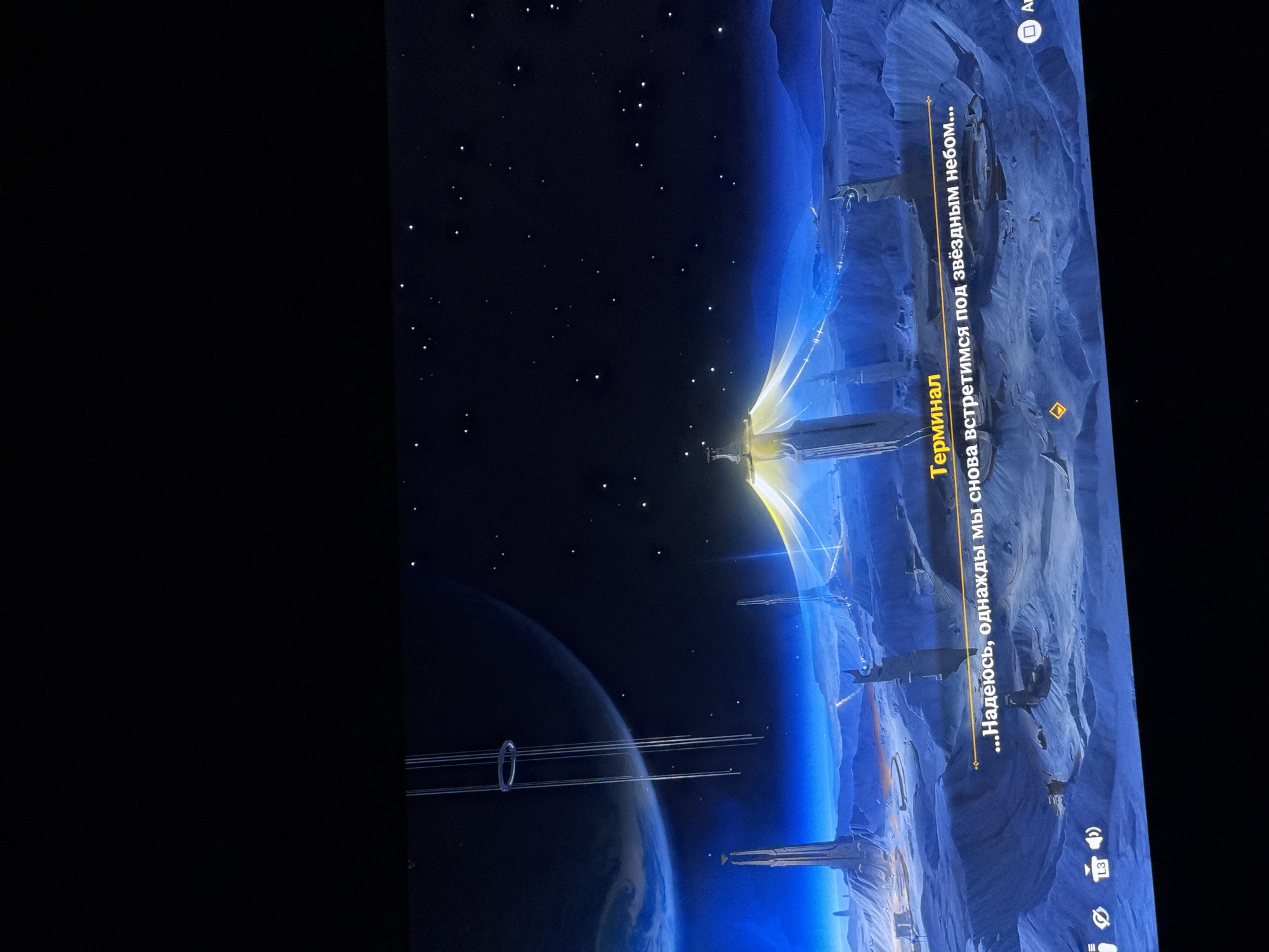 The image depicts a scene from a video game set on an alien planet. The environment is desolate and rocky, with tall, futuristic structures visible in the background. The sky is a deep blue, filled with stars, and a large planet or moon hangs in the distance. In the foreground, there are two lines of text displayed on the screen. The first line reads "Терминал" which translates to "Terminal," and the second line reads "...Надеюсь, однажды мы снова встретимся под звездным небом..." which translates to "...I hope we meet again under the starry sky...". There are also icons at the bottom left and right of the screen, suggesting user interface elements of the game. The overall impression is one of science fiction and contemplation.