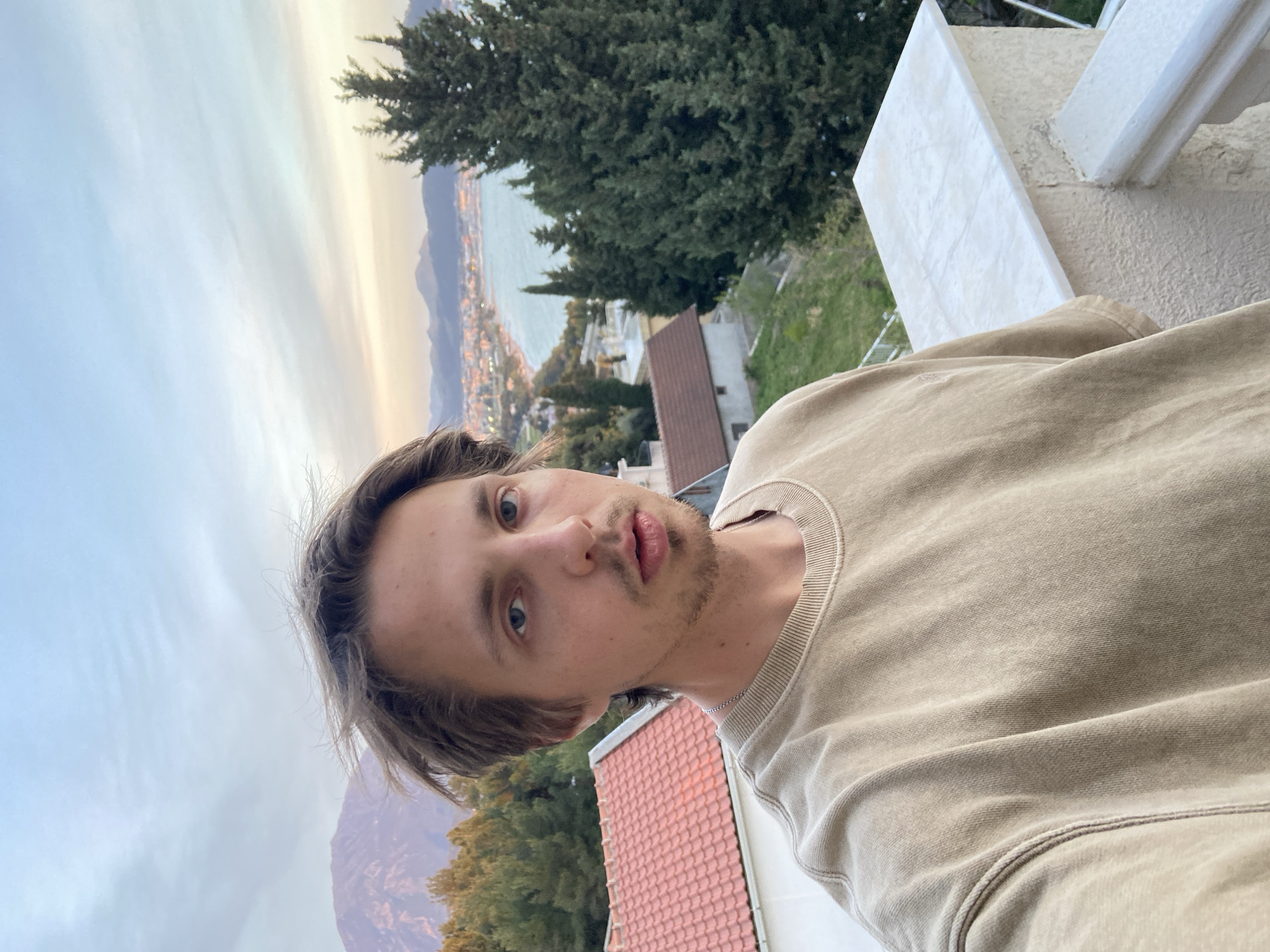 A selfie of a young man with light brown hair and a stubbled beard, wearing a tan sweatshirt, against a backdrop of Šušanj, Montenegro. The setting is a balcony or elevated area overlooking the city. In the background, there is a view of the Adriatic Sea, mountains, and the cityscape. Buildings with red tile roofs and green vegetation are visible. The sky is overcast. The man is looking directly at the camera with a slightly quizzical expression. The image is rotated approximately 90 degrees counter-clockwise.