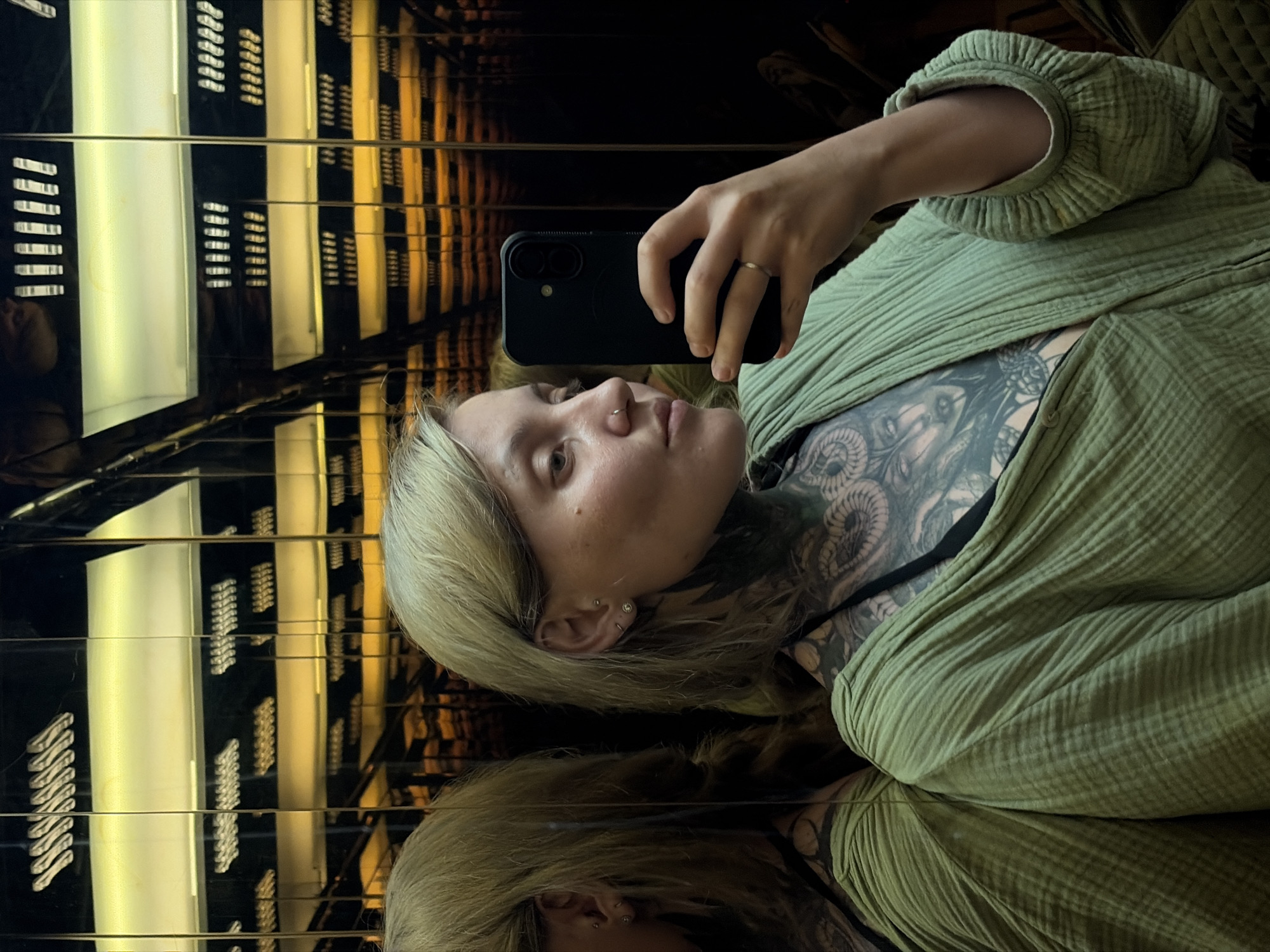 A person with blonde hair is captured in a selfie within a highly reflective, mirrored environment, likely an art installation or immersive space in Da Nang, Vietnam. The individual, appearing to be female, is positioned diagonally, looking towards the black smartphone held in their right hand. They wear a light green, textured, loose-fitting garment, which partly reveals extensive black and grey tattoos across their neck, shoulder, and upper chest, featuring intricate patterns that include what appears to be an eye and serpentine elements. Notable details on the person include a small nose ring and multiple ear piercings, as well as a simple ring on their right ring finger.
The background is dominated by the reflections of bright, rectangular light panels, creating an illusion of infinite depth. These panels alternate between warm yellow/gold and cooler white illumination, arranged in repeating rows and columns on dark, glossy surfaces. This creates a mesmerizing, grid-like pattern that extends into the distance, characteristic of a mirrored tunnel or infinity room. The person's reflection is clearly visible on the lower reflective surface. The lighting is artificial, suggesting an indoor setting. No visible text is present in the image.