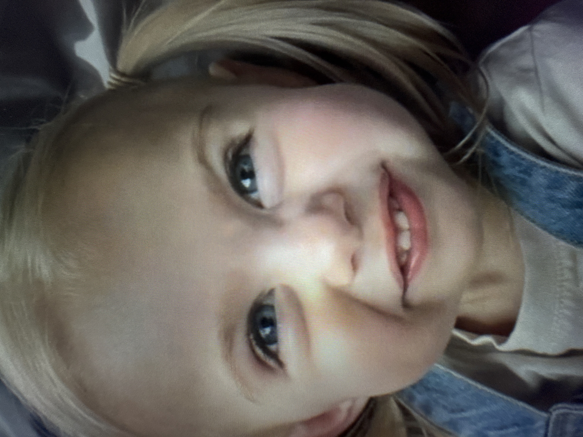 A close-up, rotated portrait image captures a young child, likely a girl, with fair skin, blue eyes, and blonde hair. Her hair appears to be styled into pigtails or two small buns on either side of her head. She is looking directly at the viewer with a bright, joyful smile, showing some teeth. Her cheeks have a natural rosy flush.
The child is wearing a light-colored, possibly off-white, top underneath blue denim overalls or a pinafore, visible on the right side of the frame. The background is soft and out of focus, suggesting a shallow depth of field or an indistinct indoor setting. The lighting is soft and even, highlighting her facial features.
A notable detail is the visible pixel grid pattern across the entire image, indicating that this photograph was taken of a digital screen or display rather than being an original digital photograph. The image is tilted approximately 90 degrees counter-clockwise, with the child's head resting on her left shoulder from the viewer's perspective. No text is visible in the image.