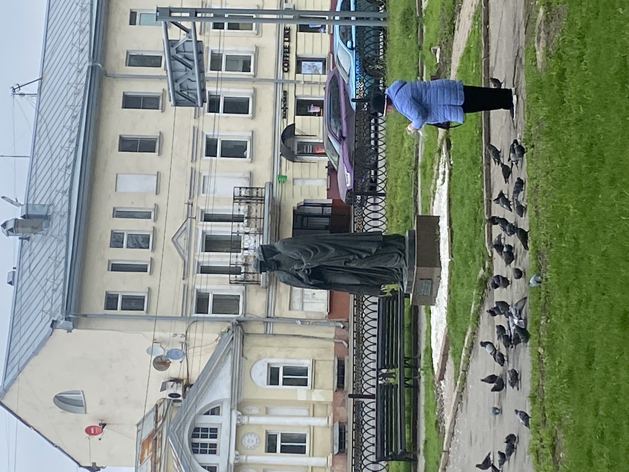 A medium, eye-level shot shows a cityscape scene with a statue, a woman feeding pigeons, buildings, and vehicles, seemingly in the city of Заволжье, Russia.
A statue is placed in front of a multi-story building with rows of windows. A black wrought-iron fence and benches surround the statue. A woman in a blue jacket and black pants stands on the right, apparently feeding a flock of pigeons on a concrete sidewalk bordered by a green grassy area.
Behind the statue, the building has a light yellow facade with symmetrically arranged rectangular windows. A coffee shop, "COFFEE LIKE", is located on the ground floor of the building. Various cars are parked along the street in front of the building.
The scene appears to be a public square or park area. The lighting is overcast, suggesting a cloudy day.