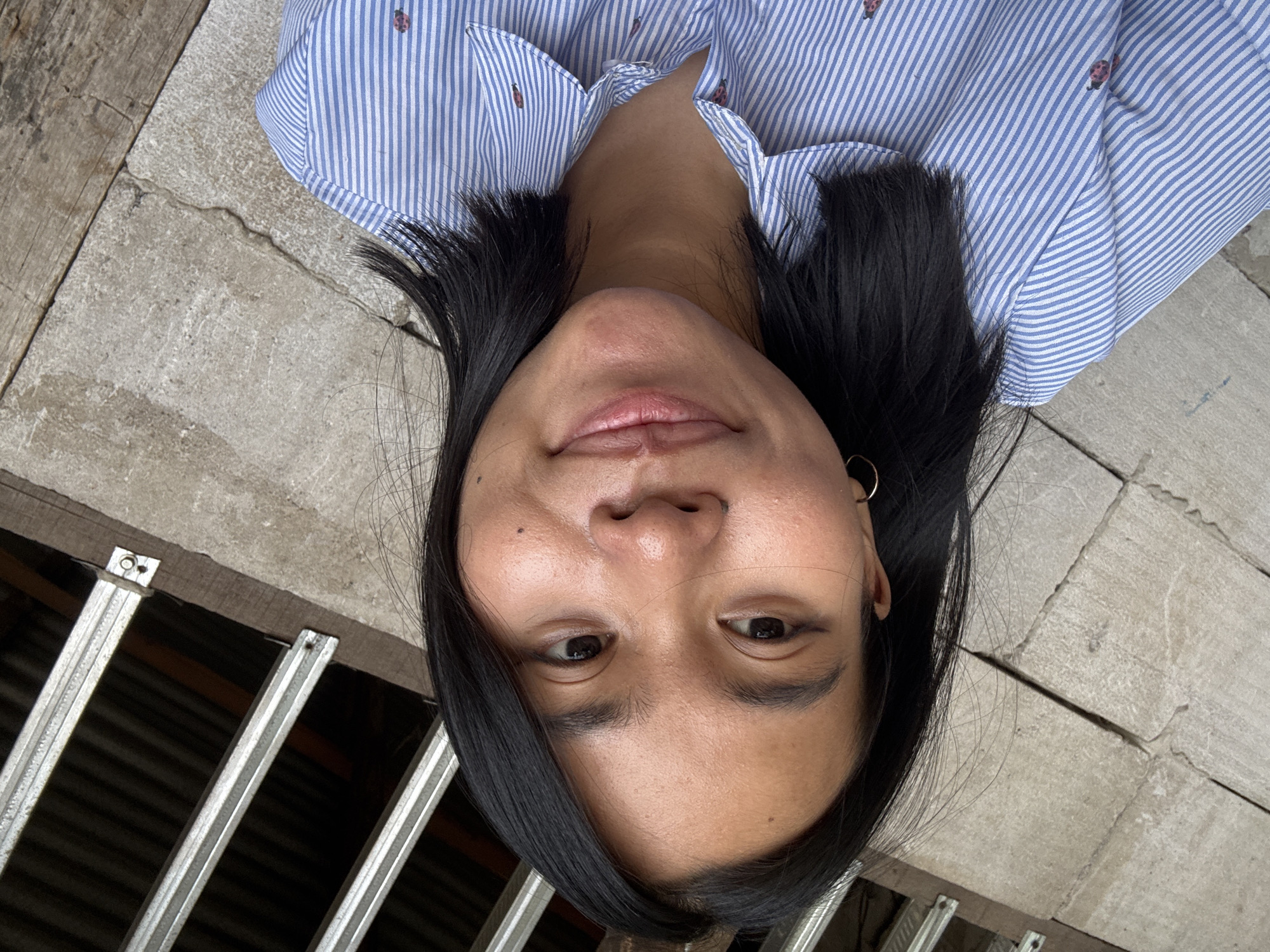 A young woman with shoulder-length black hair is taking a selfie outdoors. She is wearing a light blue and white striped button-down shirt with small ladybug patterns. Her hair is parted in the middle and frames her face. She has dark brown eyes and is smiling subtly. A small hoop earring is visible on her left ear. The background consists of rough, grey concrete walls and some metallic structures, suggesting an urban or semi-urban outdoor setting. The lighting indicates it is daytime. The scene appears to be located in East Jakarta, Indonesia.