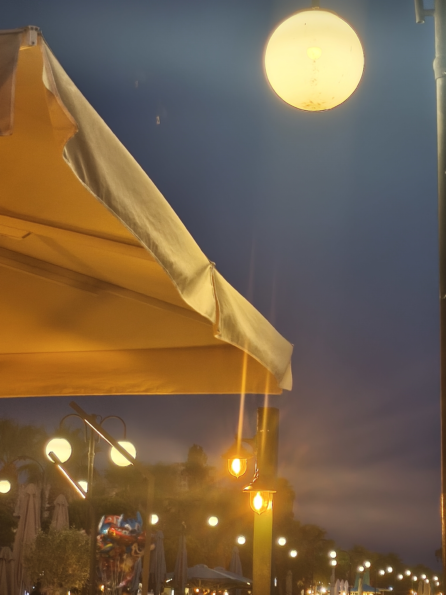 A low-angle, full-frame, eye-level shot depicts an outdoor scene at dusk in Eretria Municipal Unit, Greece. A large, yellow umbrella or canopy occupies the left and upper portions of the frame, casting a warm glow. In the upper right, a round, bright yellow streetlamp emits a strong light against a deep blue sky. A vertical pole, possibly part of a streetlight, runs along the right edge of the frame. In the middle ground, several more streetlights with glowing yellow bulbs are visible, along with closed umbrellas or beach chairs. A cluster of colorful balloons is situated in front of some of the closed umbrellas. Trees and other structures are faintly visible in the background through the hazy light. The overall atmosphere suggests a coastal or public area during evening hours.