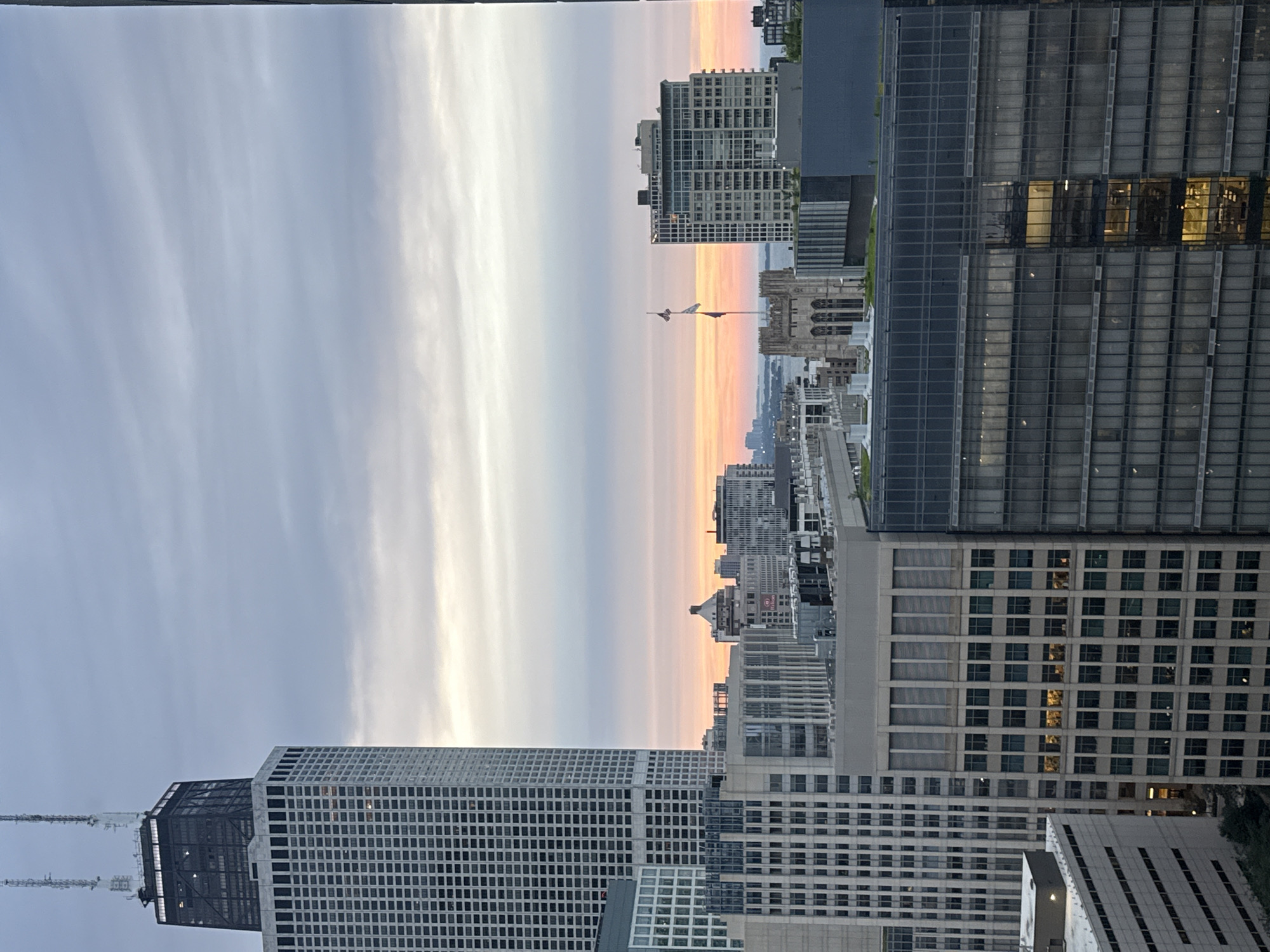 This is a photograph of the Chicago skyline taken from an elevated vantage point at dusk. The iconic John Hancock Center, a supertall skyscraper, dominates the left side of the frame. Its distinctive black top section with antennae is visible against a cloudy sky. Below it, a grid of windows stretches across the building's facade.
To the right of the John Hancock Center, a collection of mid-rise and high-rise buildings make up the urban landscape. In the middle ground, a Gothic-style building with arched windows stands out, topped with flagpoles flying flags. Beyond that, the sky exhibits a gradient of colors, transitioning from pale grey at the top to soft oranges and pinks near the horizon, indicating sunset or sunrise. A body of water, likely Lake Michigan, is faintly visible in the distance.
The foreground is filled with more buildings, many with reflective windows showing interior lights. The overall scene conveys a sense of urban density and the vastness of the city. There are no people or active interactions visible in the frame. The photograph is likely taken from another building's window or balcony, looking out over the city.