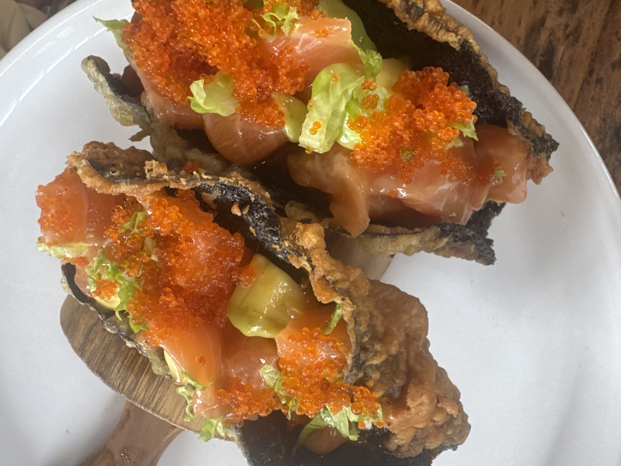 A close-up shot displays two food items, likely a fusion dish or a modern take on tacos, presented on a white plate. The tacos feature a fried, dark-colored shell, possibly seaweed or a similar ingredient, filled with raw salmon, avocado, lettuce, and a generous topping of orange fish roe. One of the tacos is slightly elevated, suggesting it is being held or propped up by a wooden utensil. The image suggests a restaurant or cafe setting, potentially in Nusa Penida, Indonesia, given the request for the location.