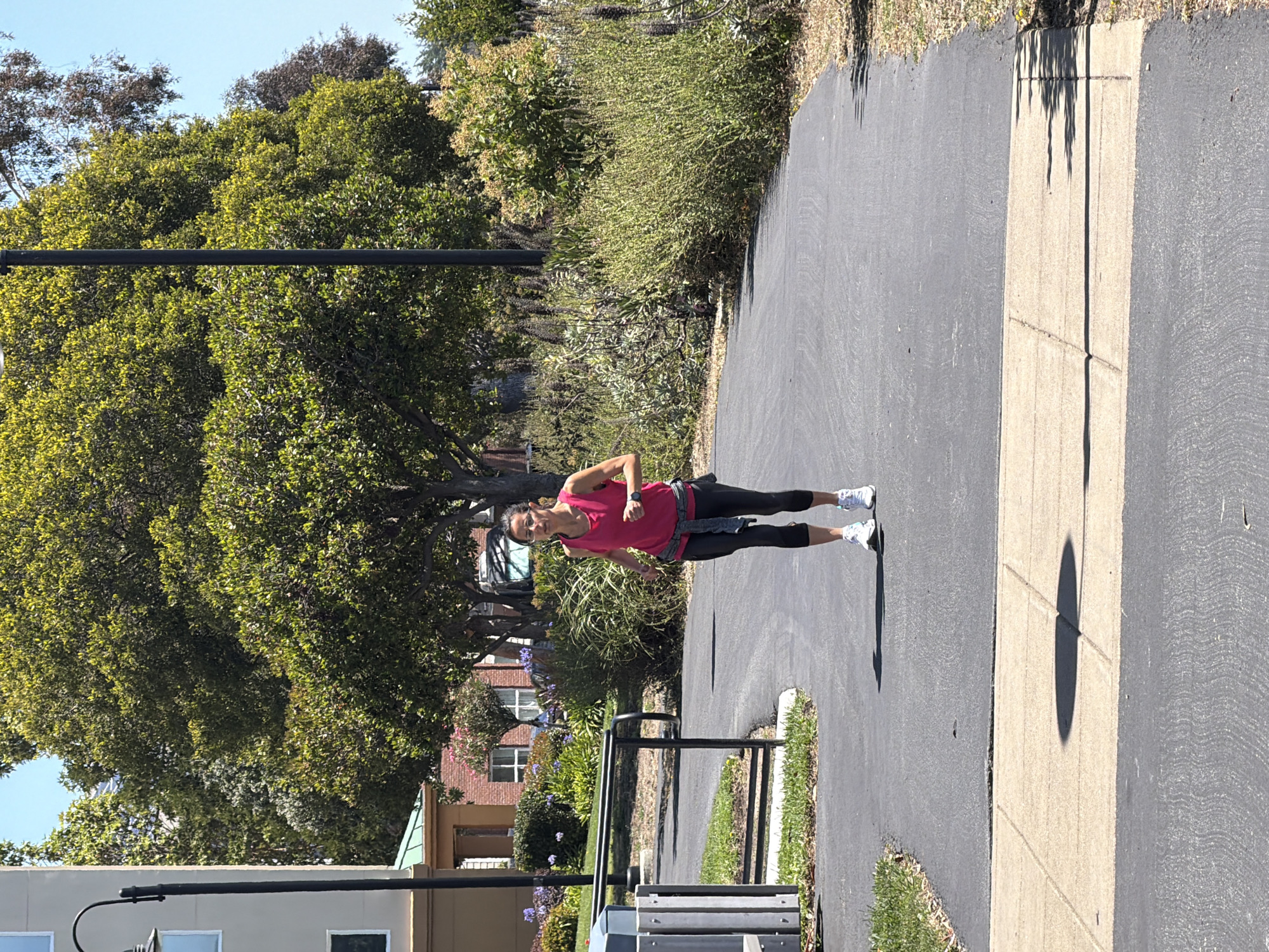 This media file is a vertical, full-body shot of a woman exercising on a paved path in what appears to be a park or residential area. The woman is wearing a bright pink tank top, black leggings, and white athletic shoes. She is positioned mid-stride, suggesting she is running or jogging. Her body is angled towards the viewer, and she is looking slightly off-camera.
The scene is outdoors, with ample greenery and trees surrounding the paved path. The path itself is a dark asphalt with a lighter-colored concrete divider running alongside it. Shadows are cast on the path, indicating it is daytime, likely mid-morning or afternoon due to the brightness and angle of the sun. The weather appears to be clear and sunny, with a bright blue sky visible in the background.
In the background, there are glimpses of buildings, one appearing to be made of red brick with white-trimmed windows, and another lighter-colored building with windows. There is also a black metal bench or rack on the side of the path. The overall atmosphere suggests a healthy and active lifestyle, with the woman enjoying an outdoor workout. There is no visible text in the image.