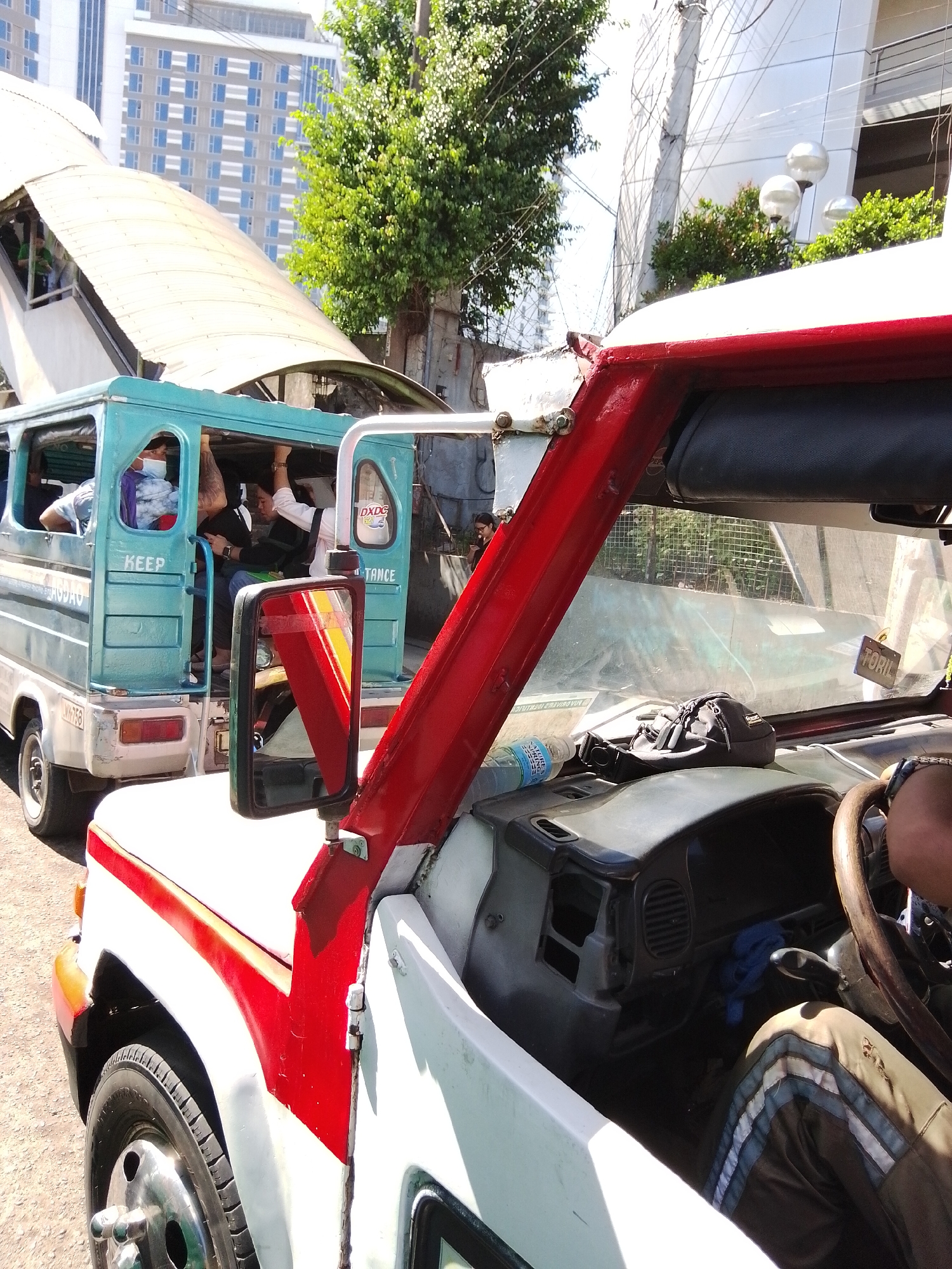 In Davao City, Philippines, on a sunny day, a jeepney with a white and red exterior is parked, with its front passenger side visible in the foreground. The jeepney's interior shows a dashboard with a steering wheel, an air vent, a blue cloth, and a grey bag on top of it. A person's legs, wearing pants with stripes, are visible near the steering wheel. A water bottle is also on the dashboard.
To the left of this jeepney, another turquoise jeepney is partially visible, also occupied by passengers. One passenger in the turquoise jeepney is wearing a face mask. Both vehicles are positioned on a paved surface, likely a street or parking area. In the background, a modern building with many windows and a green tree are prominent. Overhead, a pedestrian overpass with a white roof is also visible. The scene suggests a bustling urban environment with ongoing transportation activity.