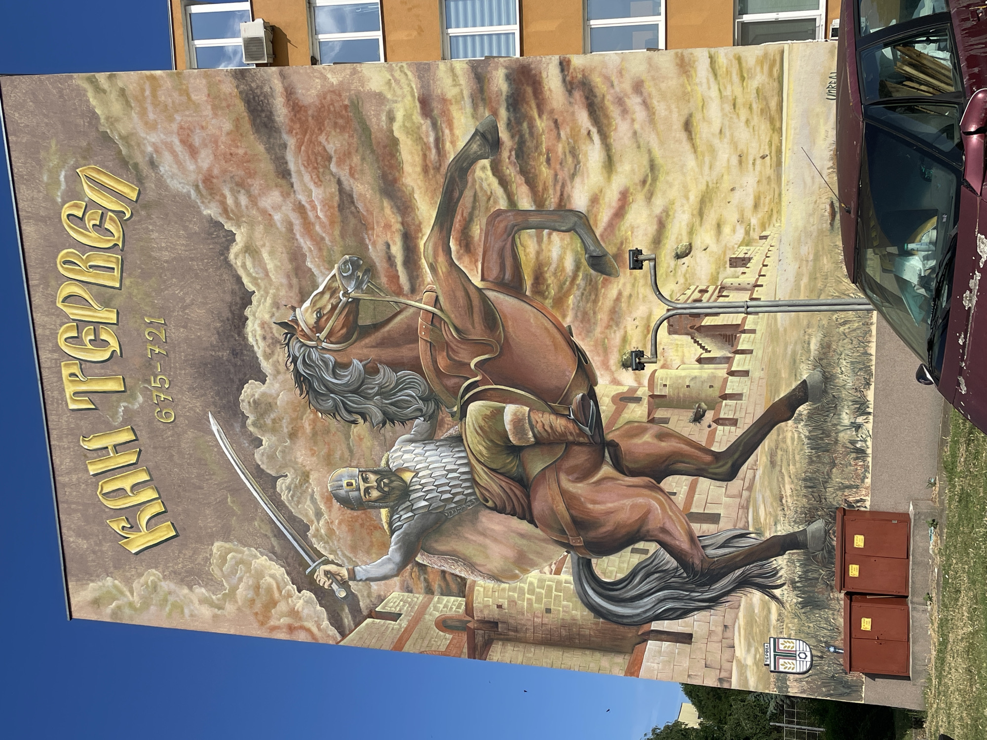 The image shows a mural painted on the side of a building, depicting a warrior on horseback in front of a castle. The mural is located in the city of Tervel, Bulgaria. The warrior is wearing armor and a helmet, and is holding a sword in his right hand. The horse is rearing up on its hind legs. The castle is made of stone and has several towers. The sky in the background is a cloudy orange color. Below the mural, there are two red electrical boxes and a burgundy car parked in front of the building. A street lamp with two lights is also visible in front of the mural. The mural is inscribed with the name "КАН ТЕРВЕЛ" and the dates "675-721". A coat of arms is visible on the lower left of the mural, with the word "ТЕРВЕЛ" below it.