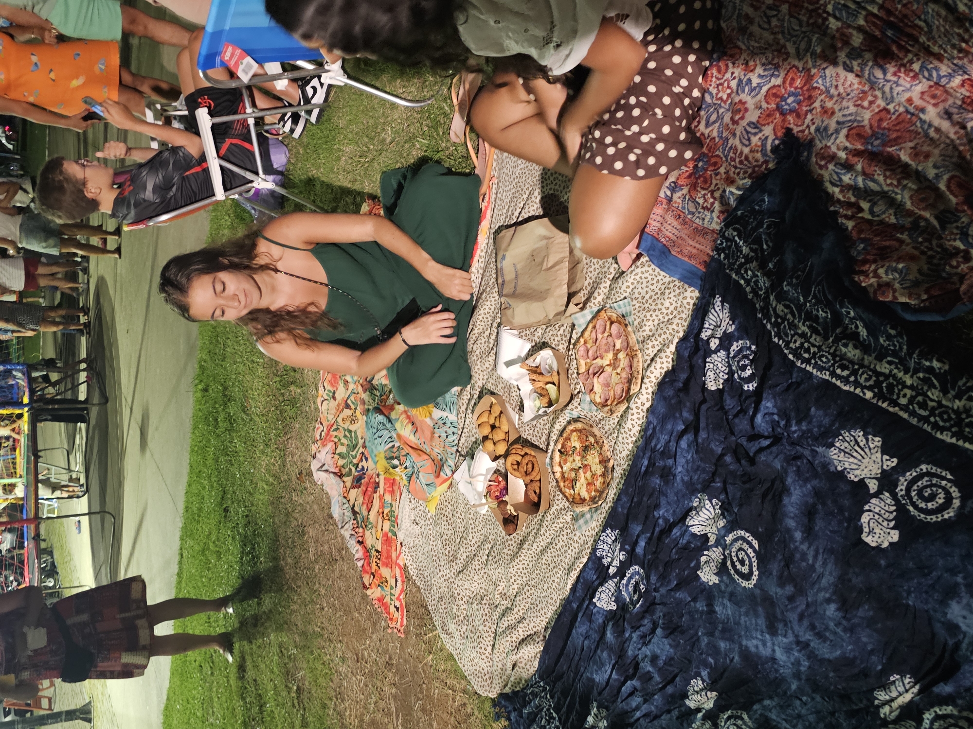 A high-angle view captures a group of people enjoying an outdoor picnic, likely in the evening. A woman in a green dress reclines on a multi-patterned blanket, surrounded by picnic fare, including pizza and fried foods in cardboard containers. Another person sits nearby, wearing a brown polka-dot skirt. The picnic blanket is spread on the grass next to a sidewalk. In the background, there are people in folding chairs and walking on the sidewalk. It seems there is a carnival going on with rides and games. Given the details of the location, this scene seems to be in Armação dos Búzios, Brazil.