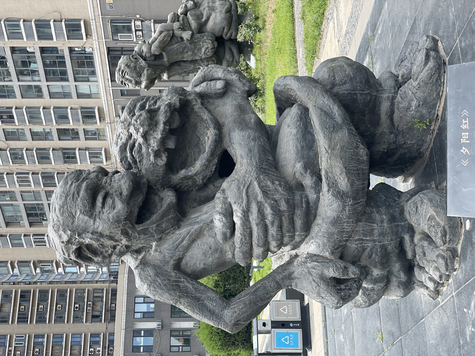 The image depicts a bronze sculpture of humans in a park-like setting. The sculpture, positioned on a black tiled base, appears to feature human figures intertwined, possibly in a struggle or embrace. Behind the sculpture, there are residential buildings with multiple windows and air conditioning units visible on the facades. Patches of grass and shrubbery are also present in the background. A trash receptacle is located near the sculpture. The image includes a placard next to the statue with Chinese characters. This scene is located in Changping District, China.