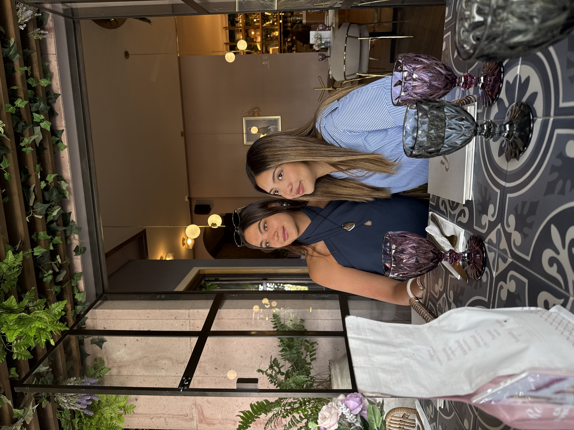 This eye-level photograph, taken in a stylish restaurant or cafe in Puebla, Mexico, features two young women seated at a table. The setting is modern and elegantly decorated with warm lighting and greenery.
On the right, a woman with long, straight brown hair wears a blue and white striped collared shirt and looks directly at the camera with a slight, pleasant smile. Next to her, on the left, another woman with dark brown hair is wearing a dark blue halter-neck top with a pendant necklace, and sunglasses are resting on her head. She also looks towards the camera with a gentle smile.
The table in front of them features a decorative gray and white patterned tile top. Two distinctively textured, faceted drinking glasses—one dark purple and one smoky gray—are placed on the table, along with a white napkin or cloth and small white booklets, possibly menus. A small floral arrangement, including lavender and light-colored roses, is visible near the foreground.
The background showcases the restaurant's ambiance, with soft, spherical artificial lights illuminating the space. Other seating areas with modern chairs and tables are visible further back, as well as framed artwork on the walls. Large glass partitions with black metal frames delineate sections of the dining area, and lush green plants like ivy and ferns are integrated into the decor, suggesting a connection to nature or an indoor garden feel. The scene appears to take place in the evening or night, given the prominent artificial lighting. Both women exhibit relaxed and happy expressions. No discernable text is clearly visible on any items.