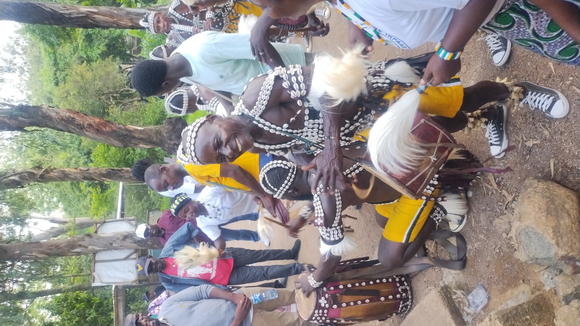 A group of people, dressed in traditional attire and modern clothing, are gathered outdoors in what appears to be a village or rural setting. The focus of the image is on a man in the foreground, wearing a headdress and ornate jewelry made of shells. He is laughing and gesturing with his hands. To his left, another man is playing a drum. Several other individuals are visible in the background, some holding what appear to be ceremonial objects. The environment consists of trees and a dirt ground, with some structures that could be simple dwellings or shelters. The scene suggests a cultural celebration or performance. The provided hints do not offer specific information to confirm the location within Jos, Nigeria.