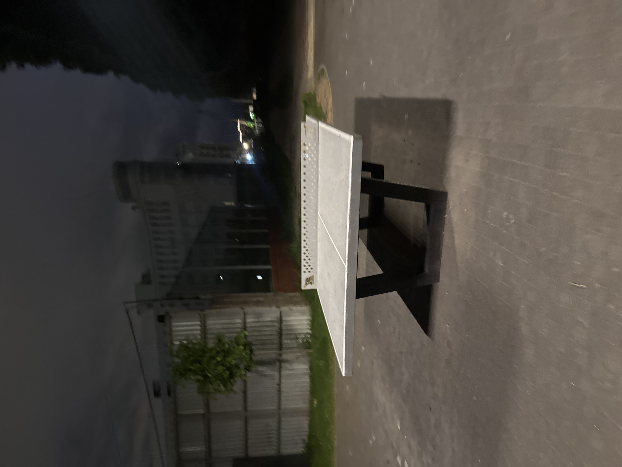 A sideways long shot depicts a table tennis table at night. The table, made of light gray material, sits on a dark gray metal frame. Its placement is on a concrete surface, with a green grass strip and a vertical fence visible along the left edge. The table is near a canal or body of water, and across the water, the reflections of a large building with many windows and streetlights are visible. This location is in Saint Petersburg, Russia, based on the architecture and canal characteristics. No people are present in the scene.